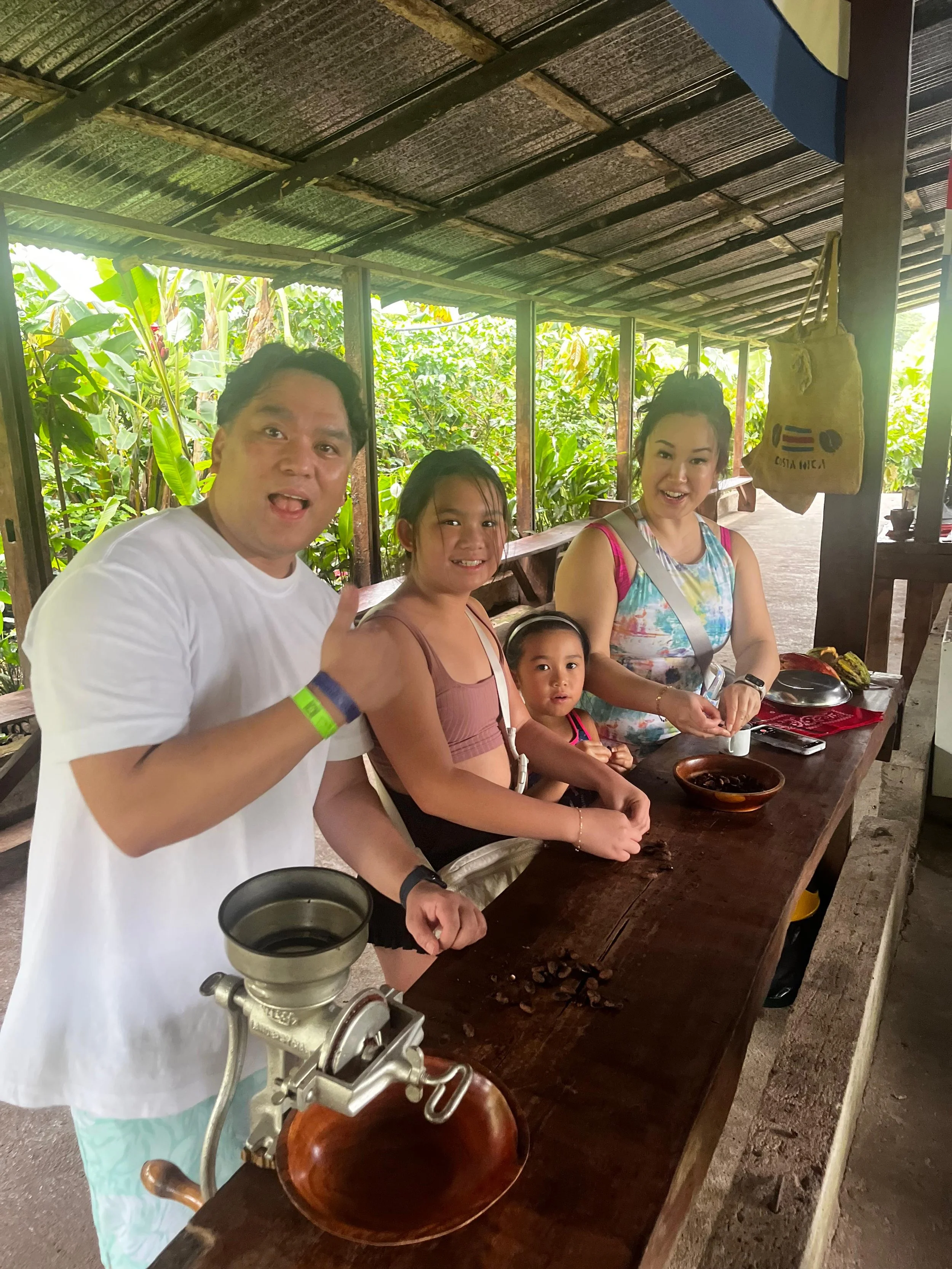 A family of four standing at a wooden counter with bowls and a meat grinder, inside a rustic open-air structure with lush green plants in the background. Liberia Airport Shuttle. Tours in Costa Rica.