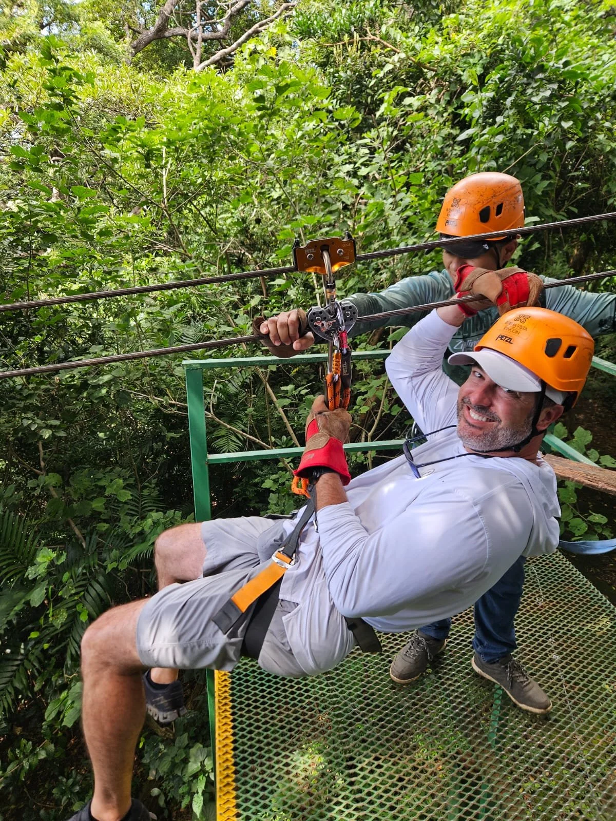 Two men wearing orange safety helmets and harnesses are on a metal platform, securing a safety line in a lush green forest. Liberia Airport Shuttle. Tours in Costa Rica.