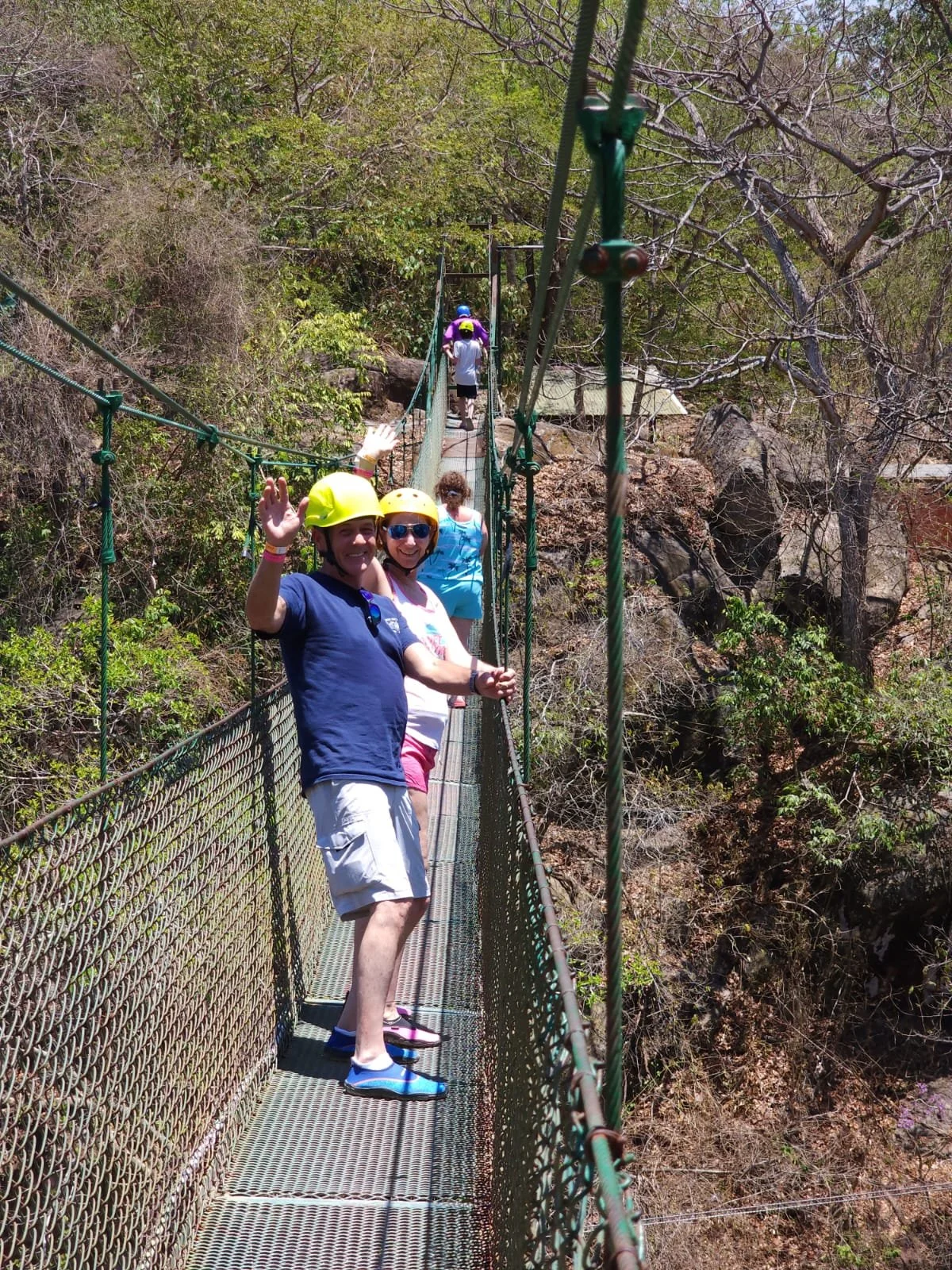 People crossing a suspension bridge in a wooded area, wearing helmets and casual clothes, enjoying an outdoor adventure. Liberia Airport Shuttle. Tours in Costa Rica.