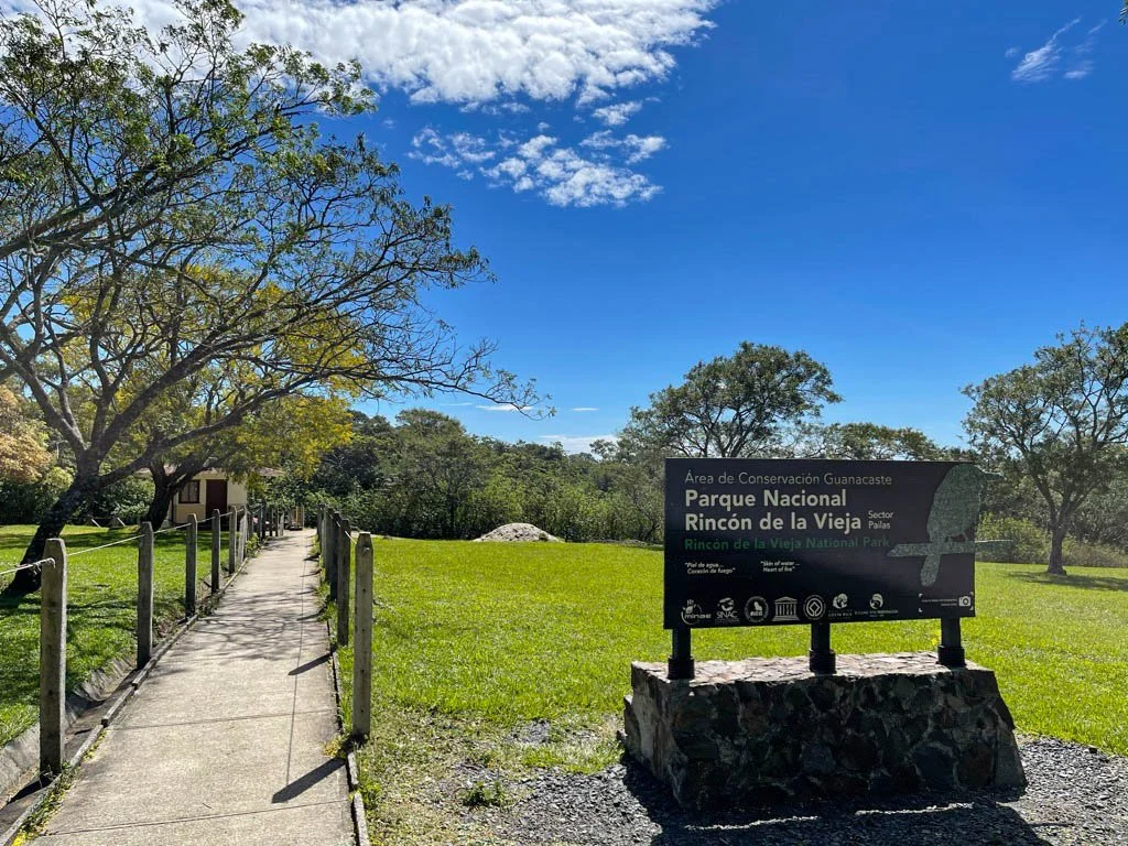 Pathway leading into a lush green park with trees and a sign for Rincón de la Vieja National Park in Costa Rica under a blue sky with scattered clouds. Liberia Airport Shuttle. Tours in Costa Rica.