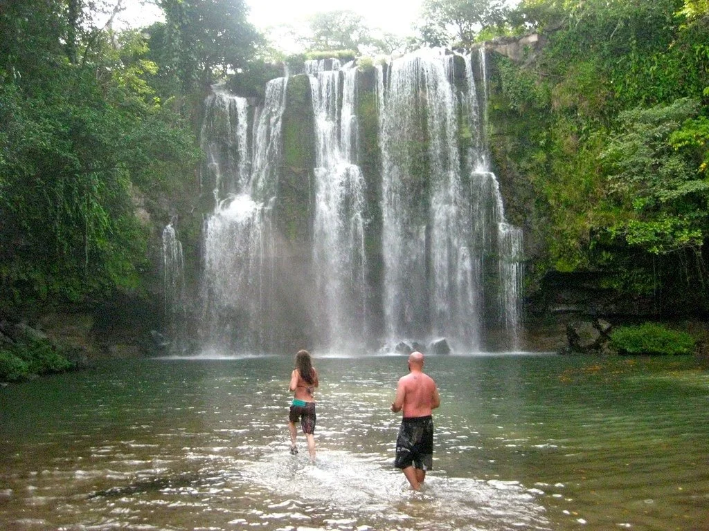 Two people, a woman and a man, stand in a shallow pond facing a large waterfall surrounded by green trees. Liberia Airport Shuttle. Tours in Costa Rica.