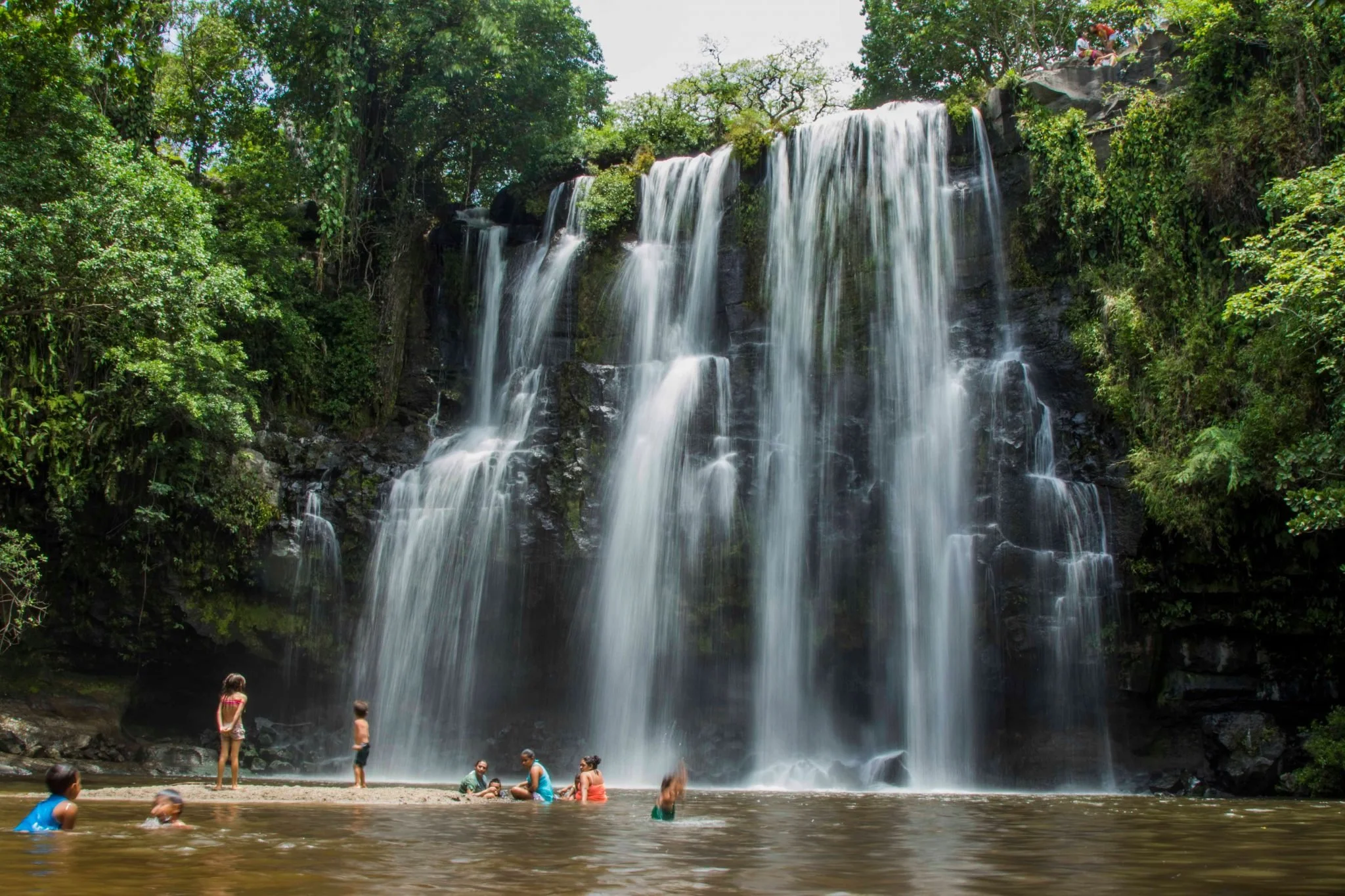 People swimming and playing near a large waterfall surrounded by lush green trees. Liberia Airport Shuttle. Tours in Costa Rica.