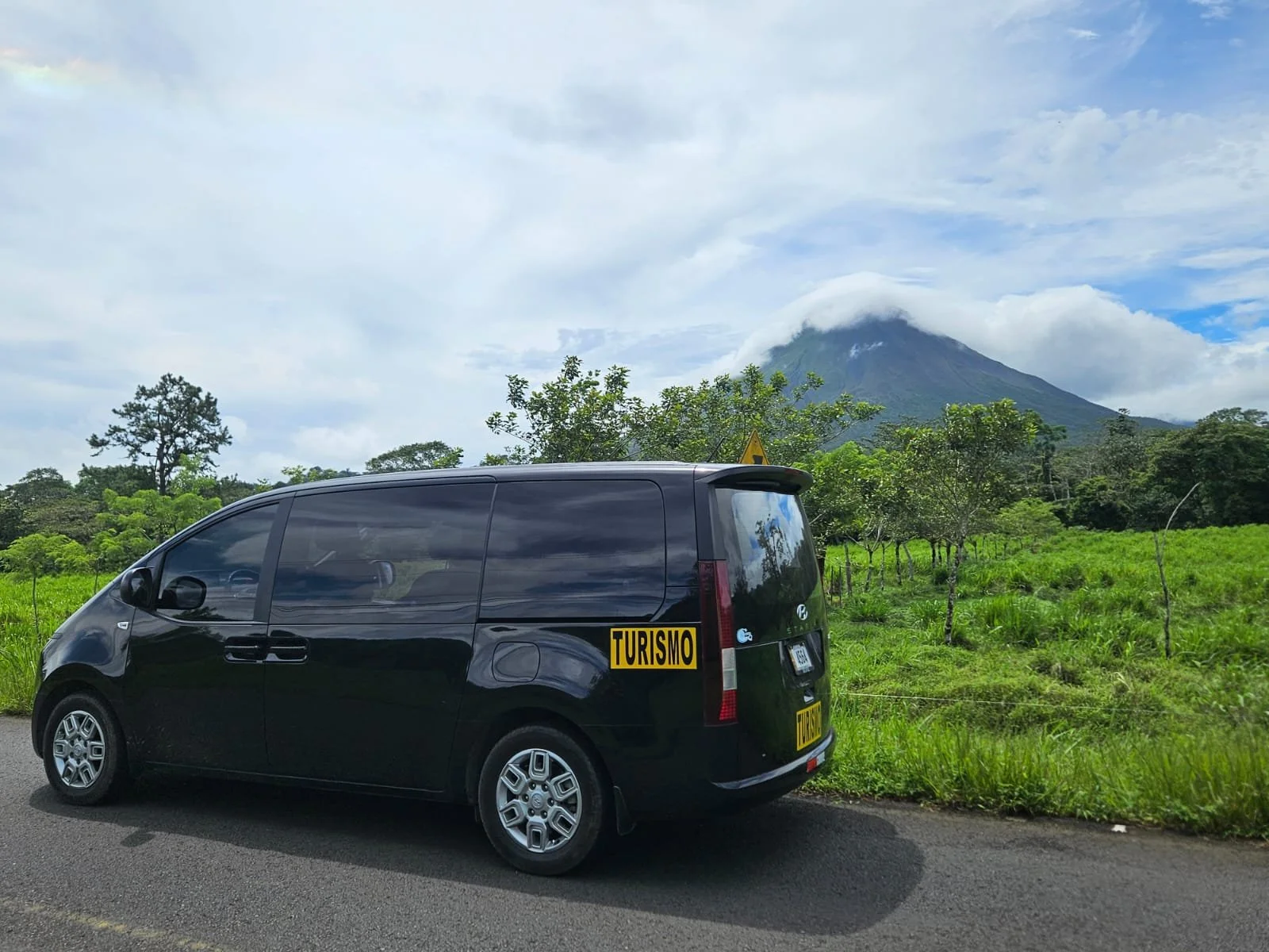 Black van parked on the side of a road with lush green fields and trees, with a mountain in the background partly covered by clouds. Liberia Airport Shuttle. Tours in Costa Rica.