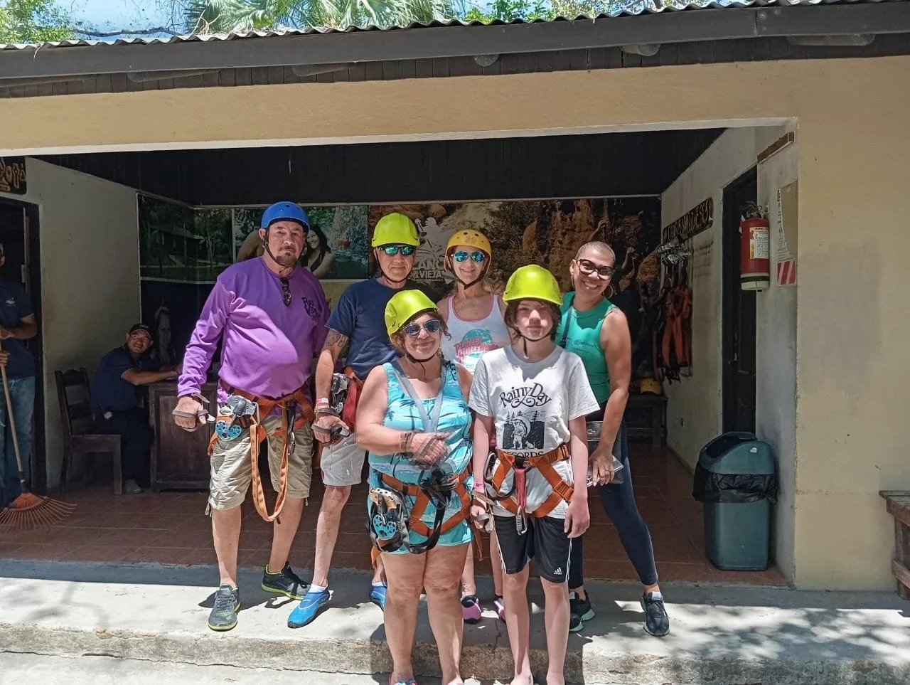 Group of six people wearing helmets and safety harnesses, standing in front of a facility, likely for outdoor adventure activities, with two additional individuals in the background. Liberia Airport Shuttle. Tours in Costa Rica.
