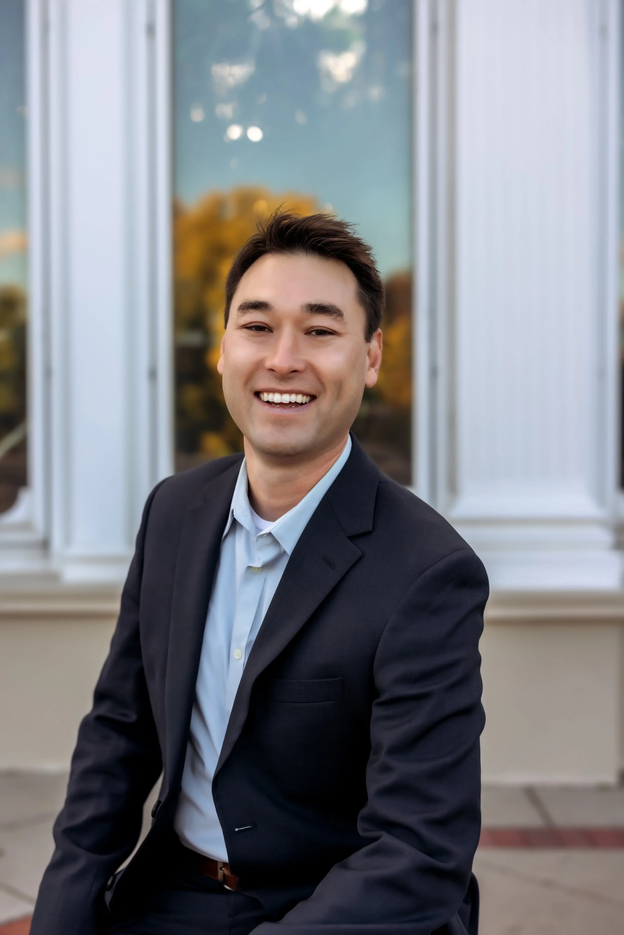 A young man with dark hair, smiling, dressed in a navy blue blazer and light blue shirt, sitting outdoors in front of a building with large windows and white columns during fall.
