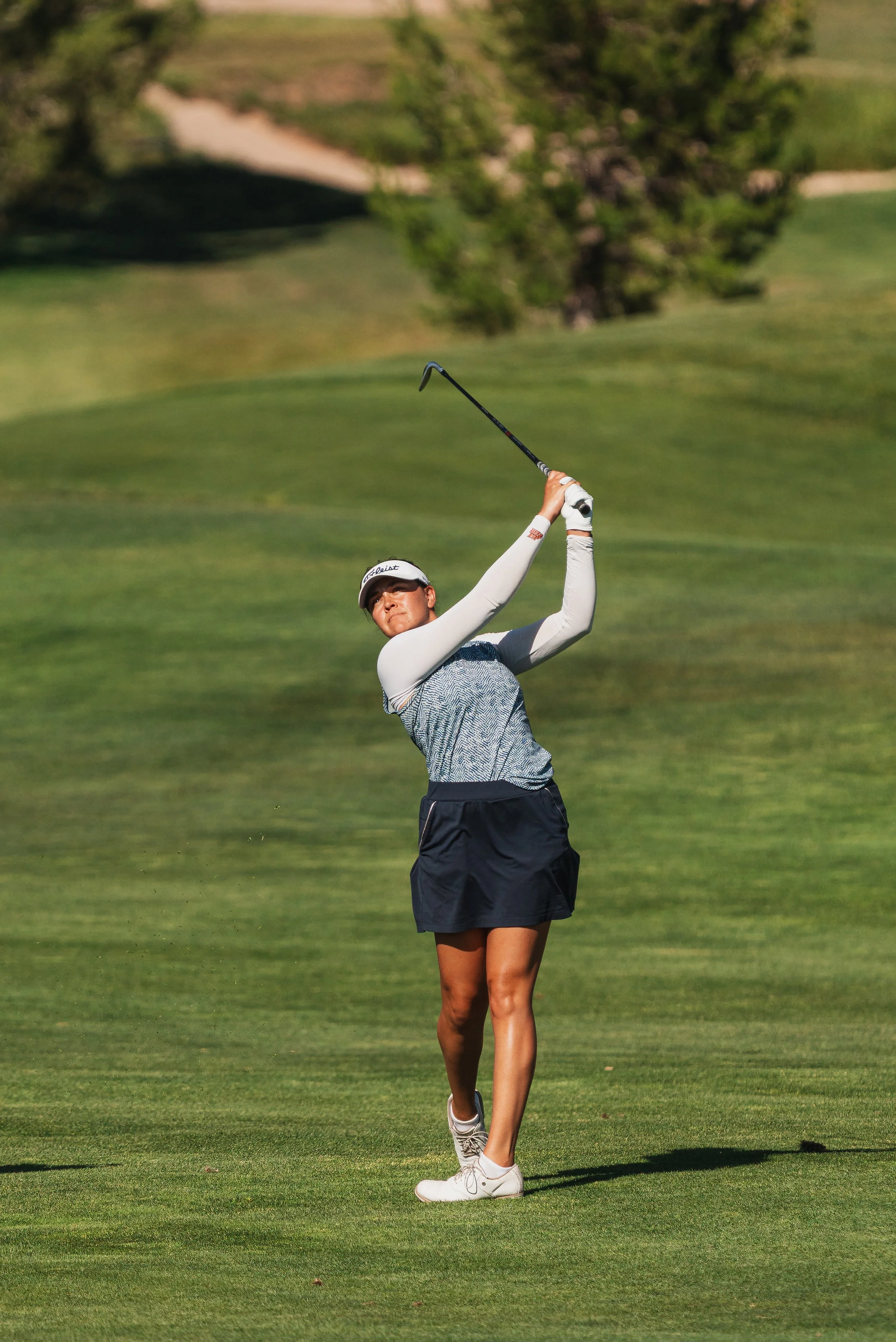 A woman in a white golf skirt and shirt swings a golf club on a lush, green golf course surrounded by trees.