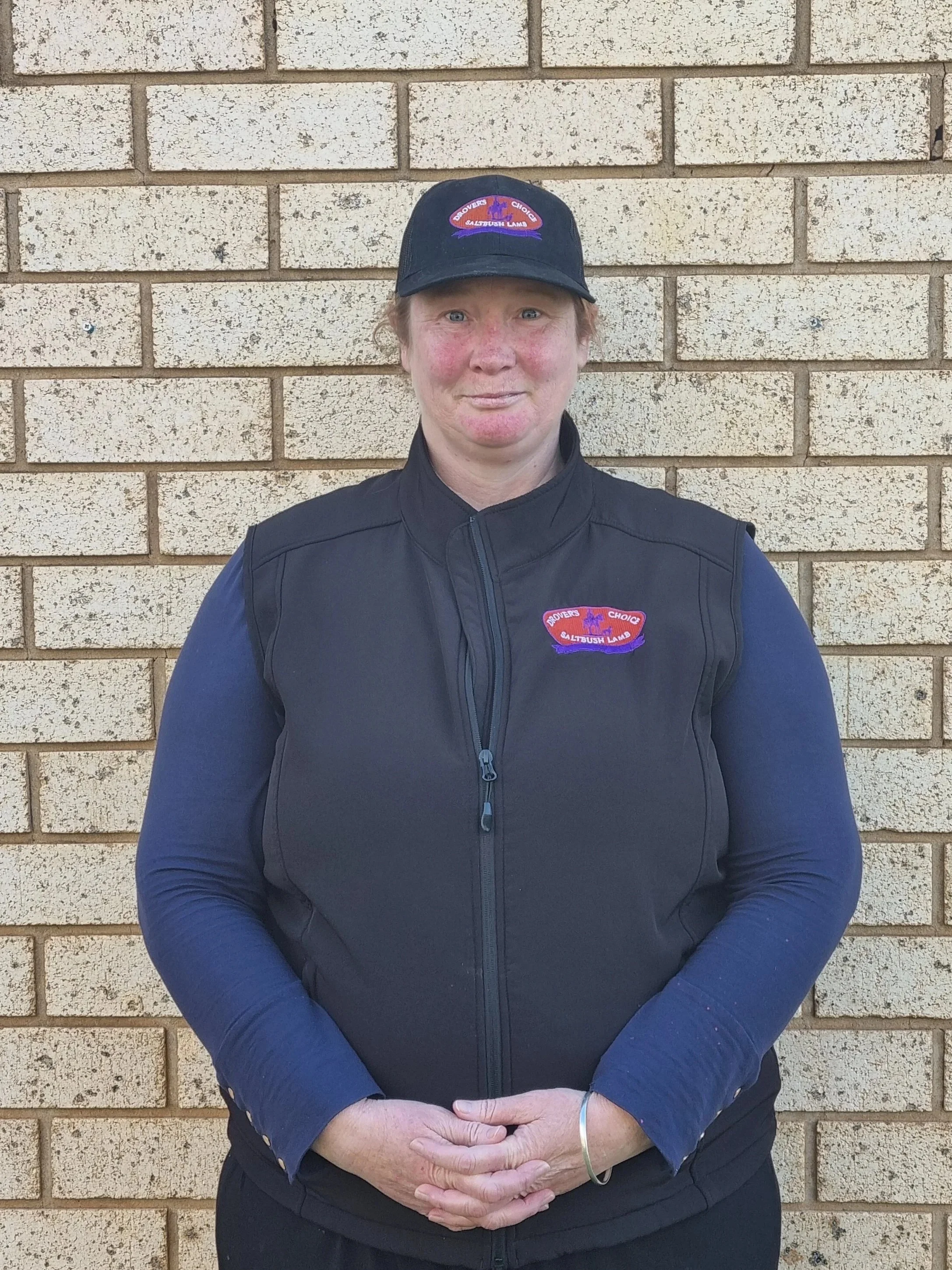 A woman wearing a black vest and matching cap standing against a brick wall, with a slight smile and hands clasped in front.