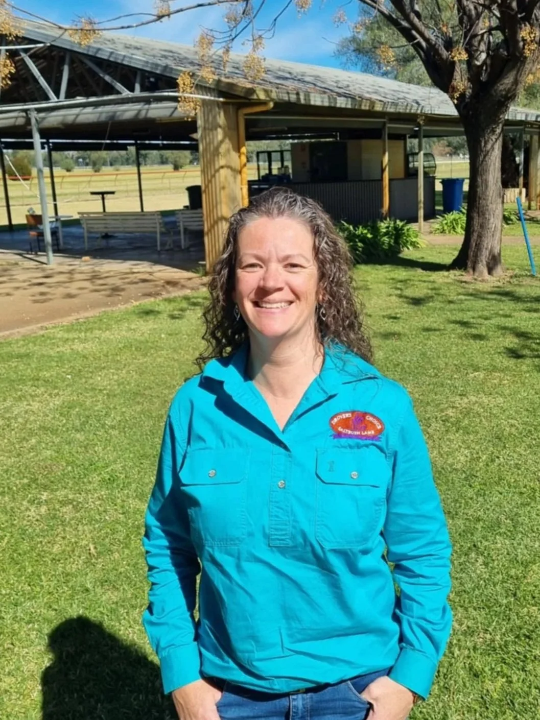 Elaine, A smiling woman with curly hair wearing a bright blue jacket standing outside on grass, with a pavilion and trees in the background under a clear blue sky.