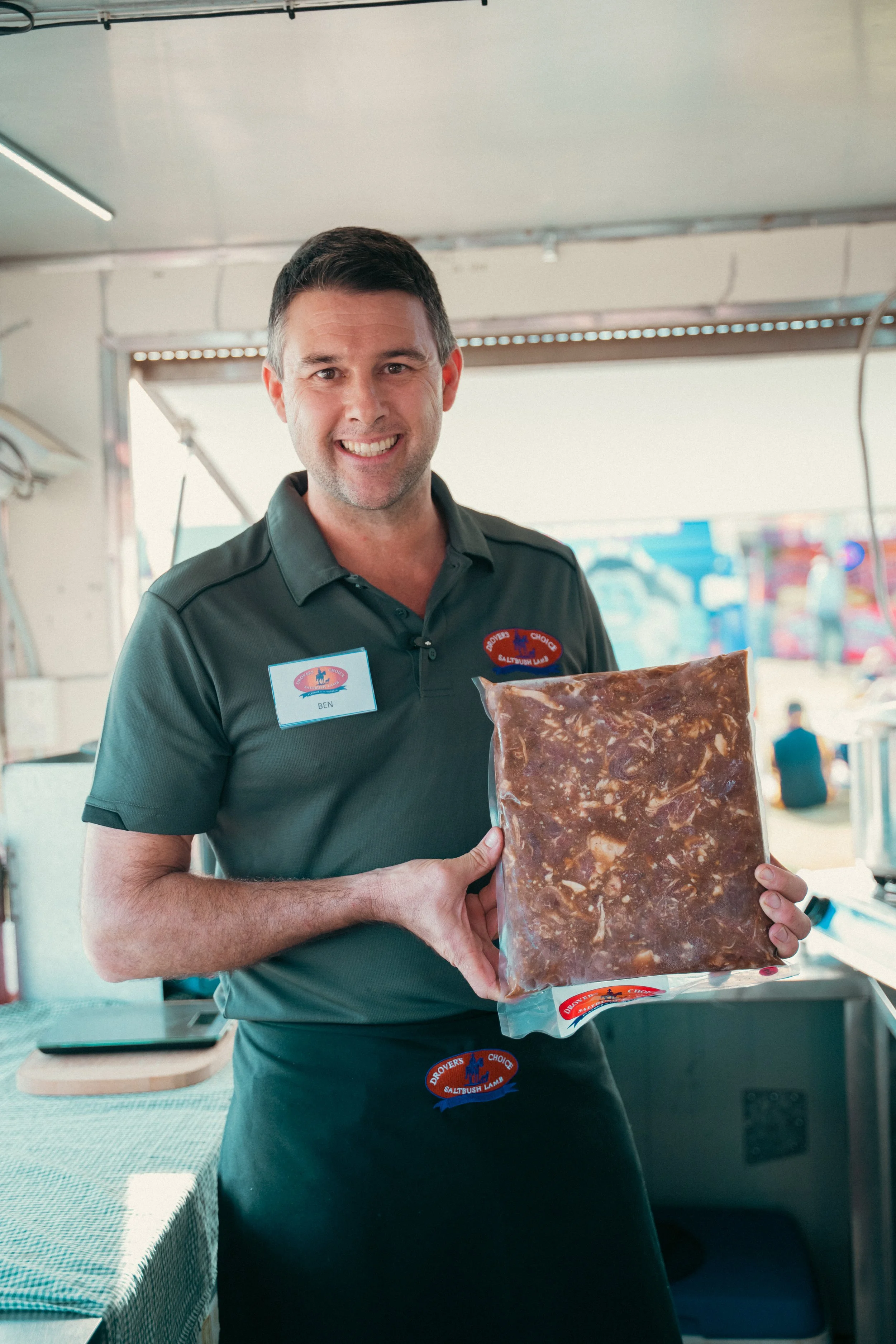 Saltbush Lamb owner in a dark green polo shirt and apron holding a large package of Saltbush lamb and gravy, at an outdoor agricultural show or festival