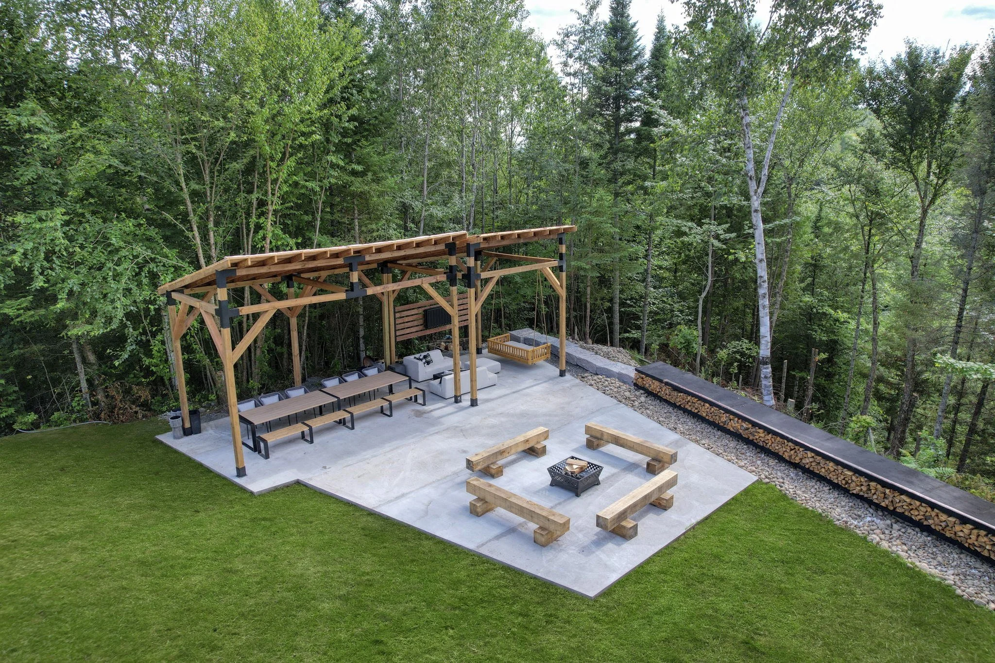 Espace extérieur avec pergola en bois, meubles de jardin, espace pour feu de camp, entouré de forêt et d'un mur de bois de chauffage.