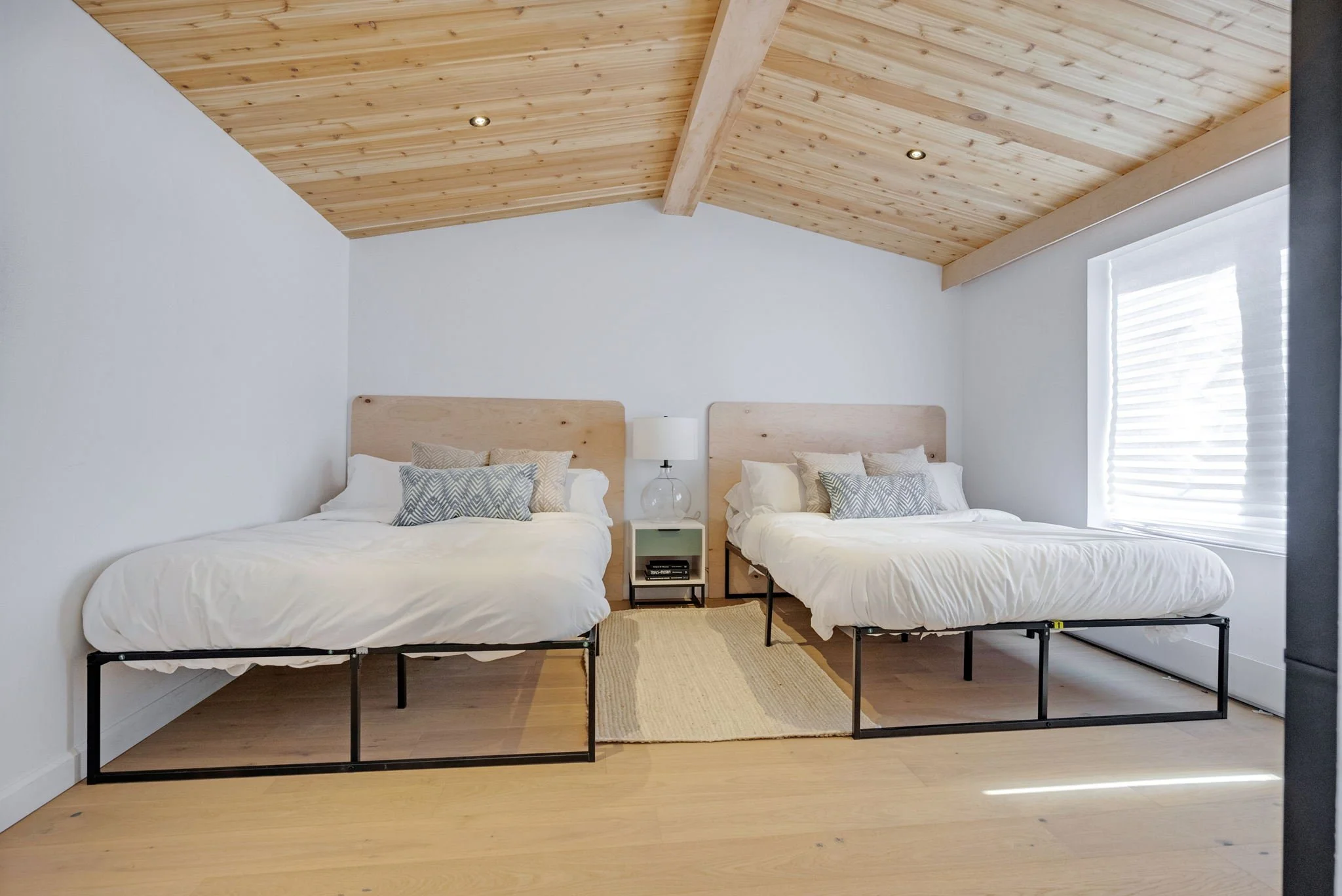 Chambre avec deux lits doubles à cadre en métal noir, tête de lit en bois clair, sur tapis beige, avec table de nuit, lampe et livres, sous un plafond en bois avec deux spots et une grande fenêtre avec stores blancs.