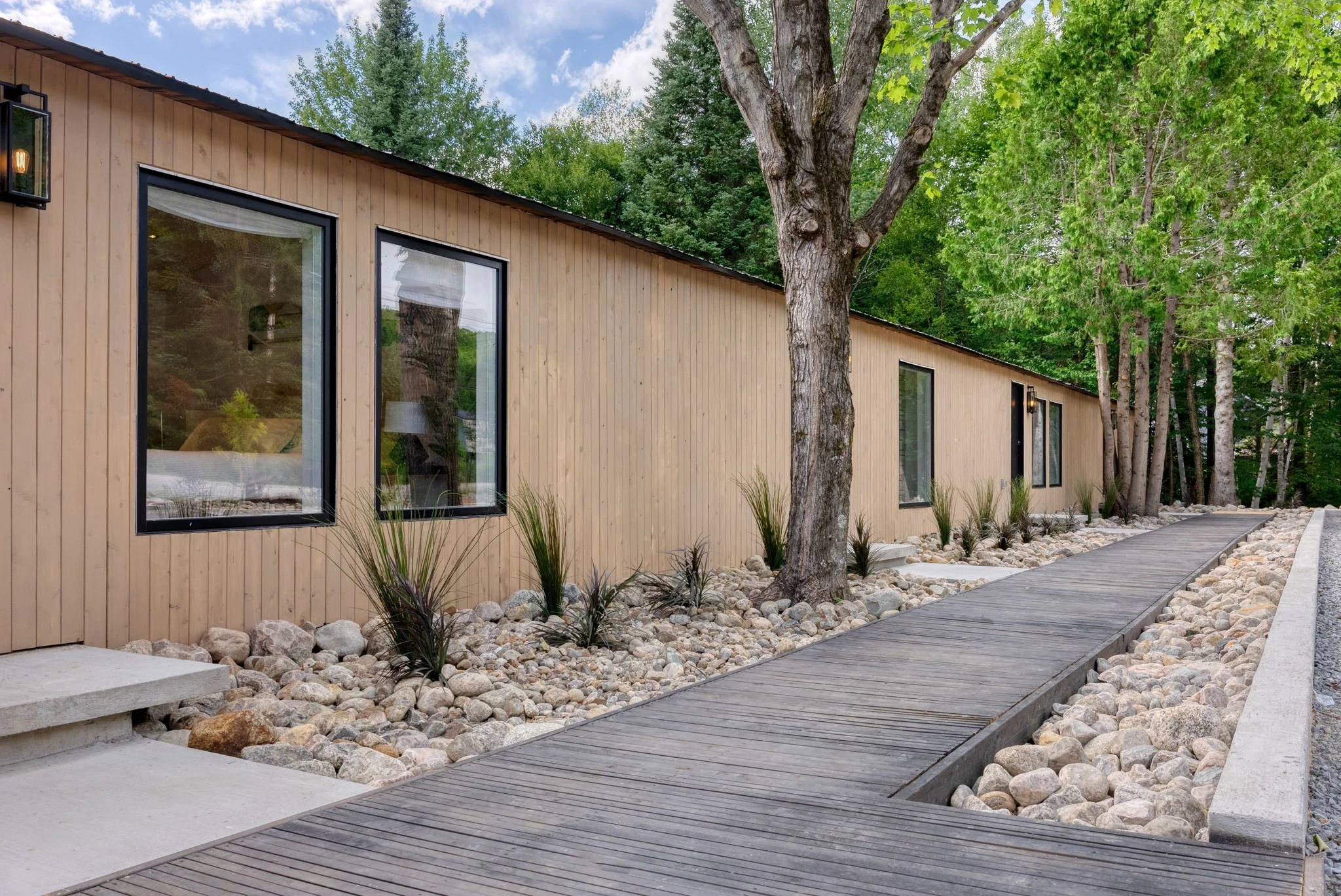 Une longue maison moderne en bois avec des fenêtres rectangulaires, entourée d'arbres et de pierres, avec un chemin en bois menant à l'entrée.