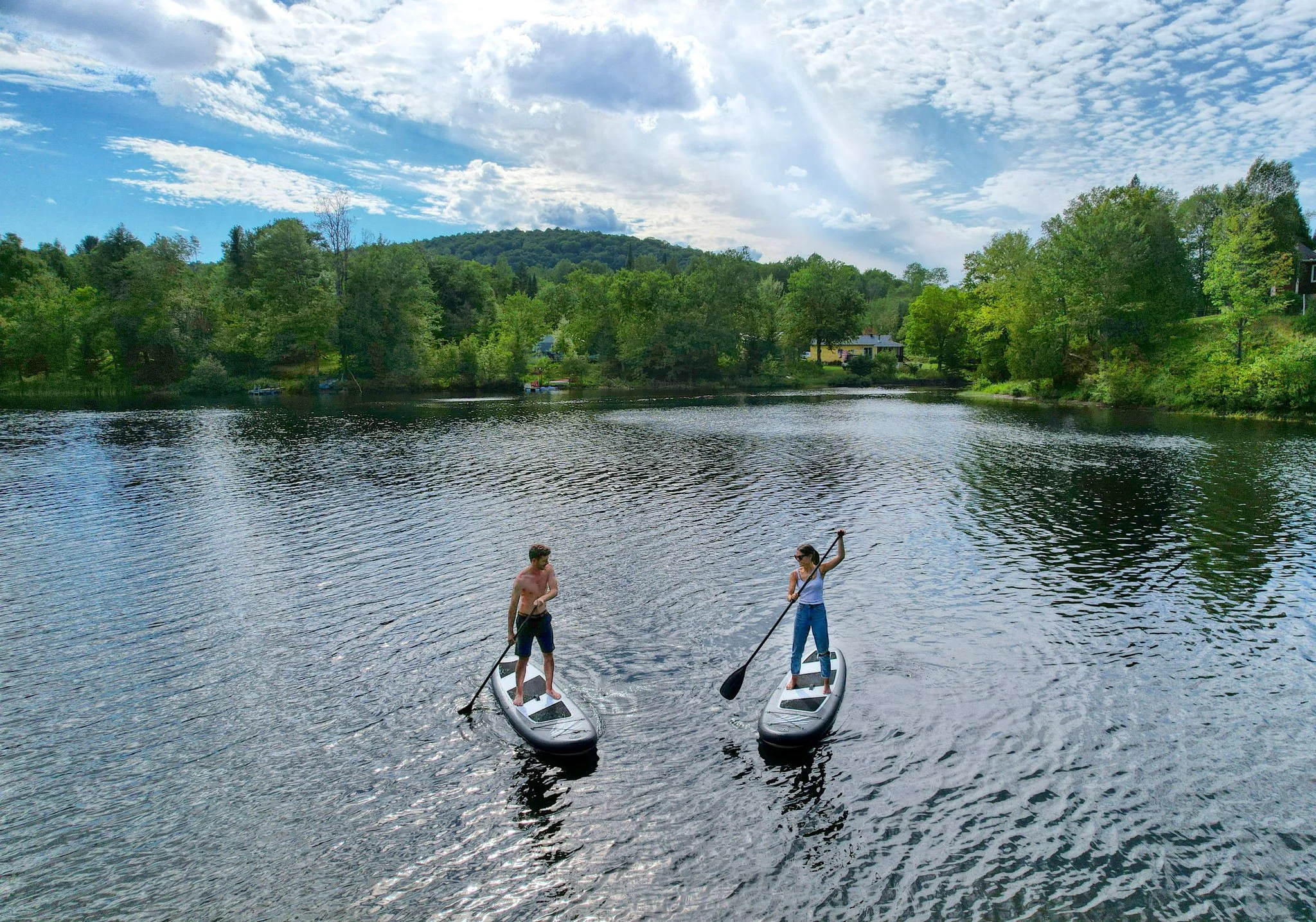 Deux personnes font du paddle sur un lac entouré d'arbres et de collines sous un ciel partiellement nuageux.