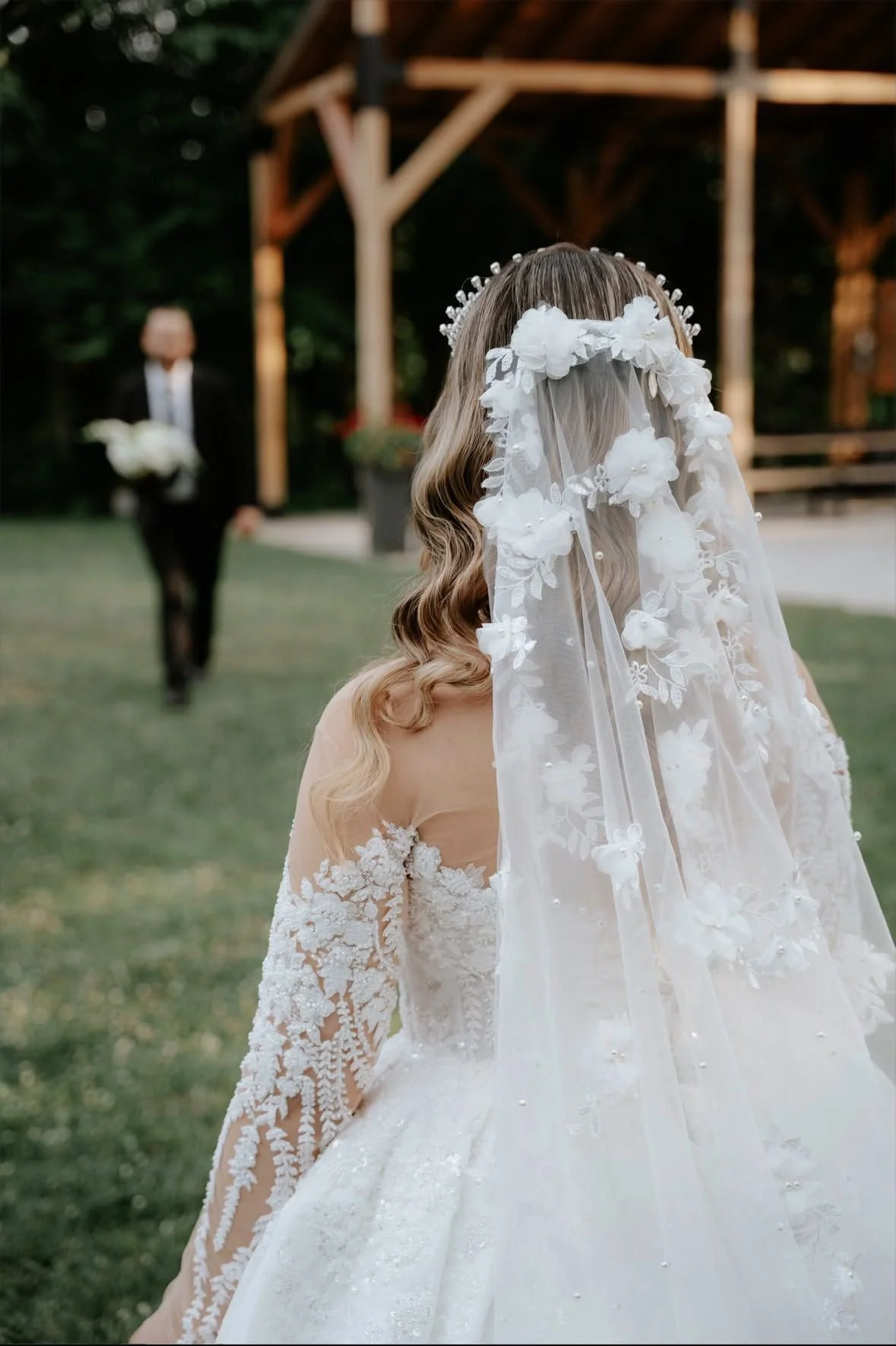 Une mariée vue de dos avec une voile ornée de fleurs et un arrière-plan de mariage en extérieur, avec un homme en costume noir approchant.