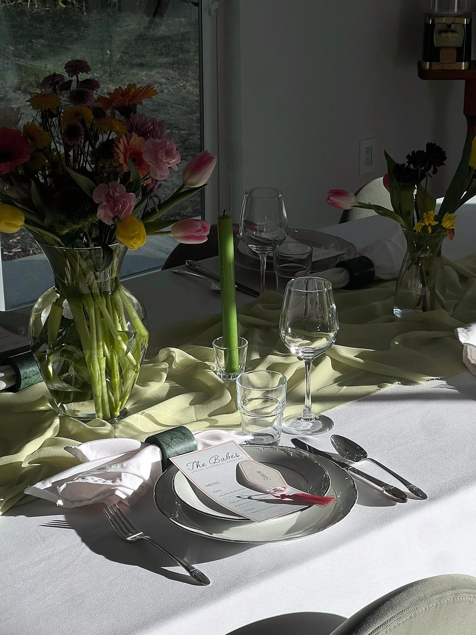 Une table de dîner élégante avec des vases contenant des fleurs, des verres à vin, des assiettes et des couverts, décorée avec un galon en tissu jaune pâle et une bougie verte au centre.