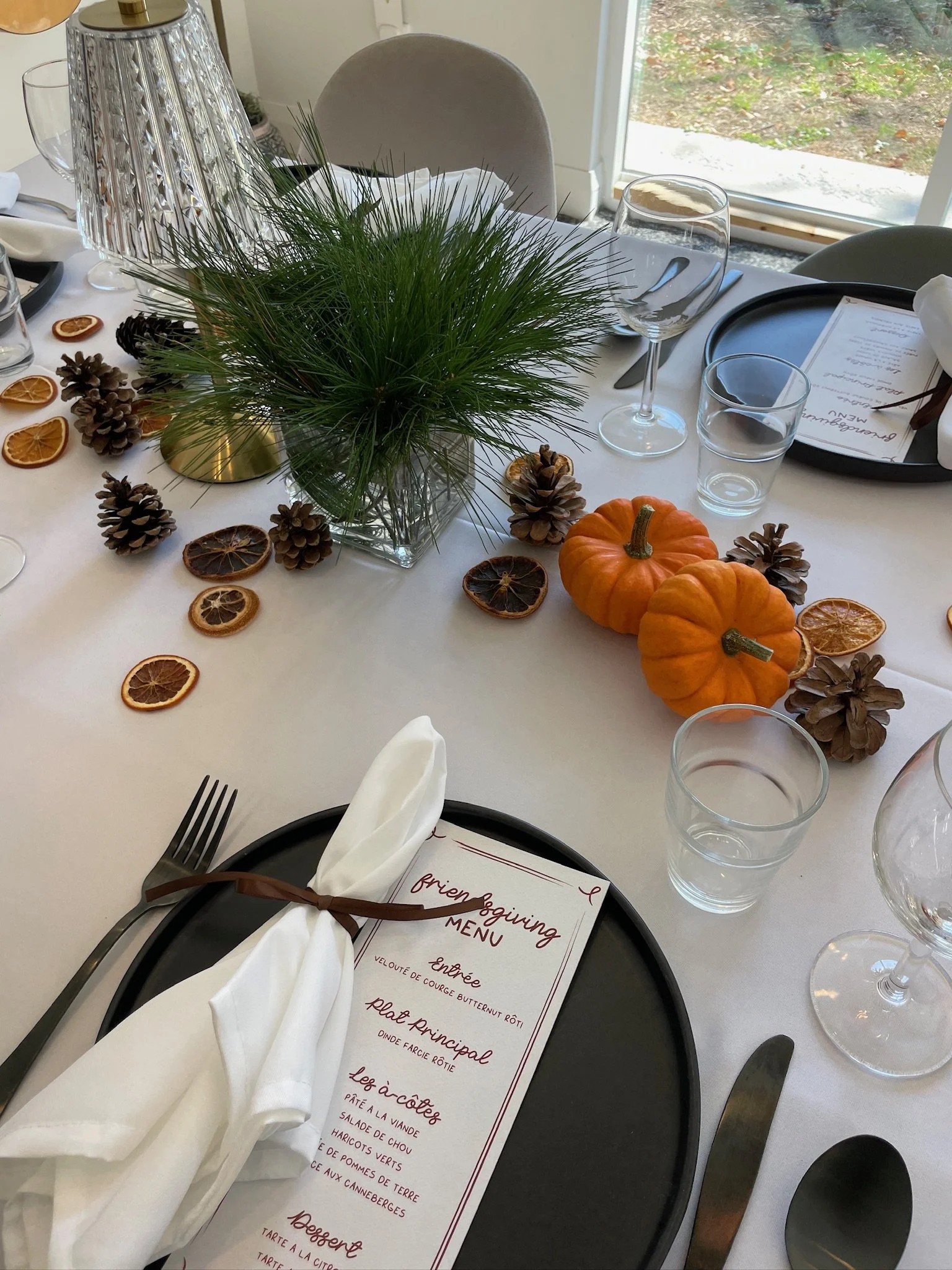Une table de dîner décorée avec un centre de table composé de branches de pin, de pommes de pin et de citrouilles miniatures. La table est prête pour un repas, avec des verres, des assiettes,noeuds en ruban blanc et un menu écrit en français.