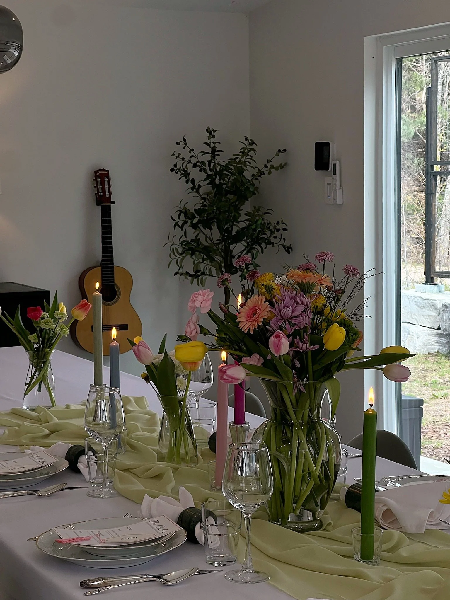 Table de fête avec des fleurs colorées, bougies allumées, assiettes, verres et couverts, dans une pièce lumineuse avec une guitare sur le mur à gauche et une fenêtre donnant sur un jardin.