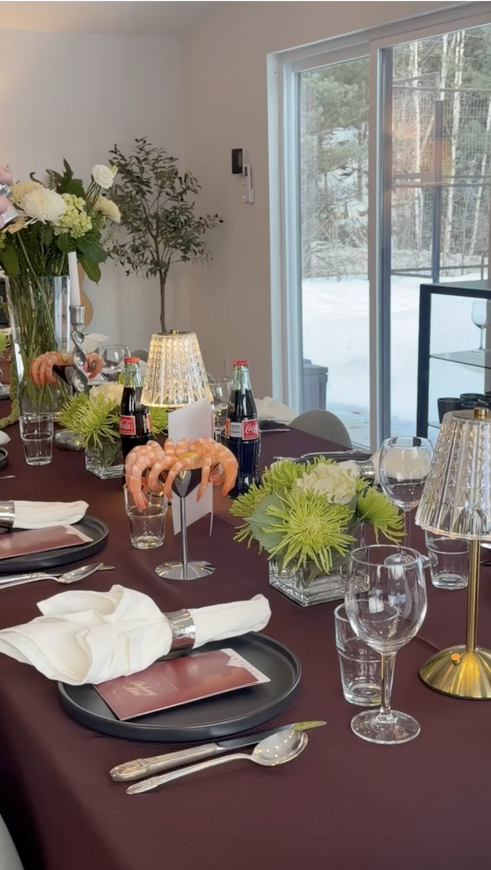 Table de dîner décorée avec des fleurs, des lampes, des verres à vin, des bouteilles de Coca-Cola, et des plats, dans une pièce avec une fenêtre donnant sur la neige extérieure.