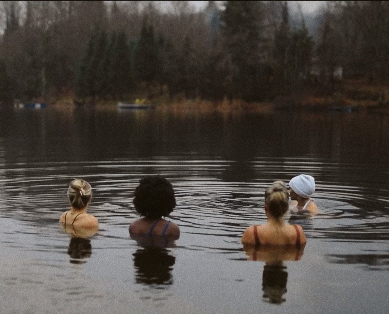 Quatre femmes nagent dans un lac, avec des arbres en arrière-plan, lors d'une journée nuageuse ou en soirée.
