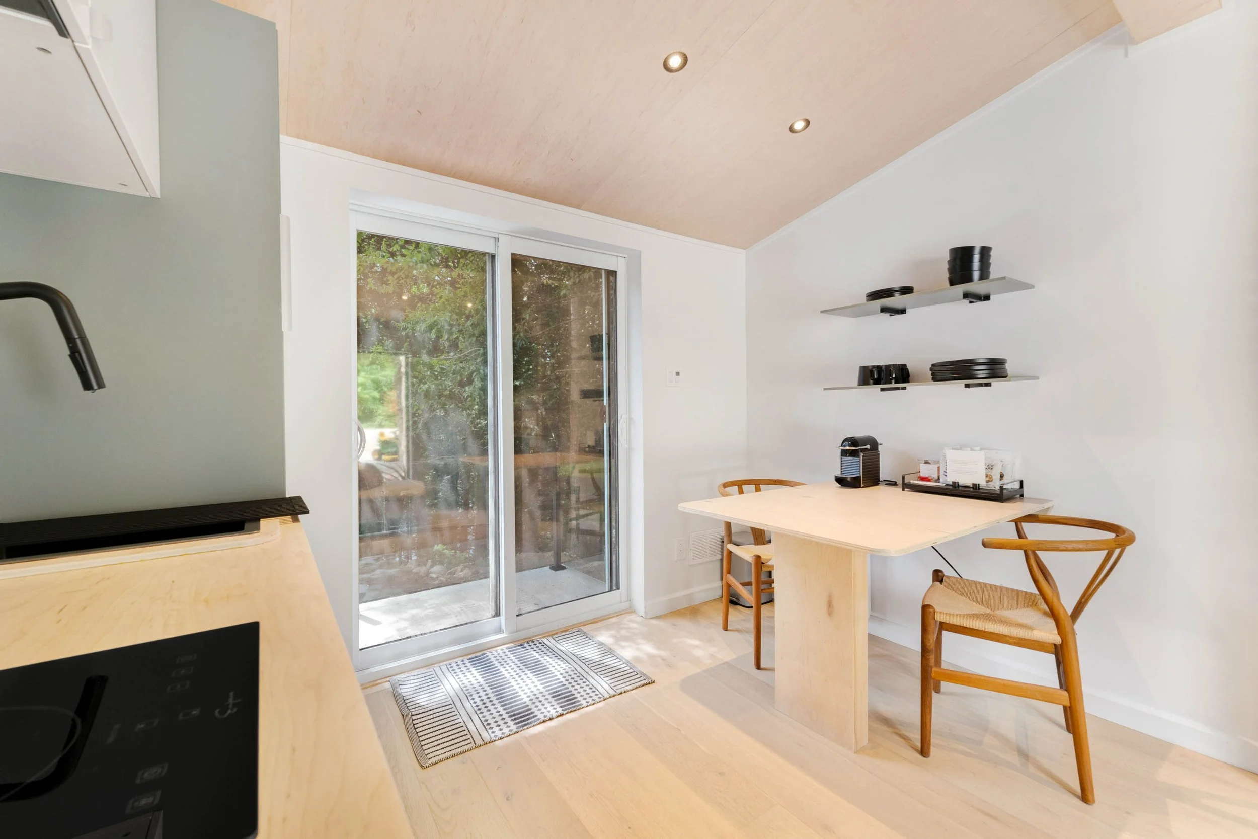 Chambre avec deux lits doubles à cadre en métal noir, tête de lit en bois clair, sur tapis beige, avec table de nuit, lampe et livres, sous un plafond en bois avec deux spots et une grande fenêtre avec stores blancs.