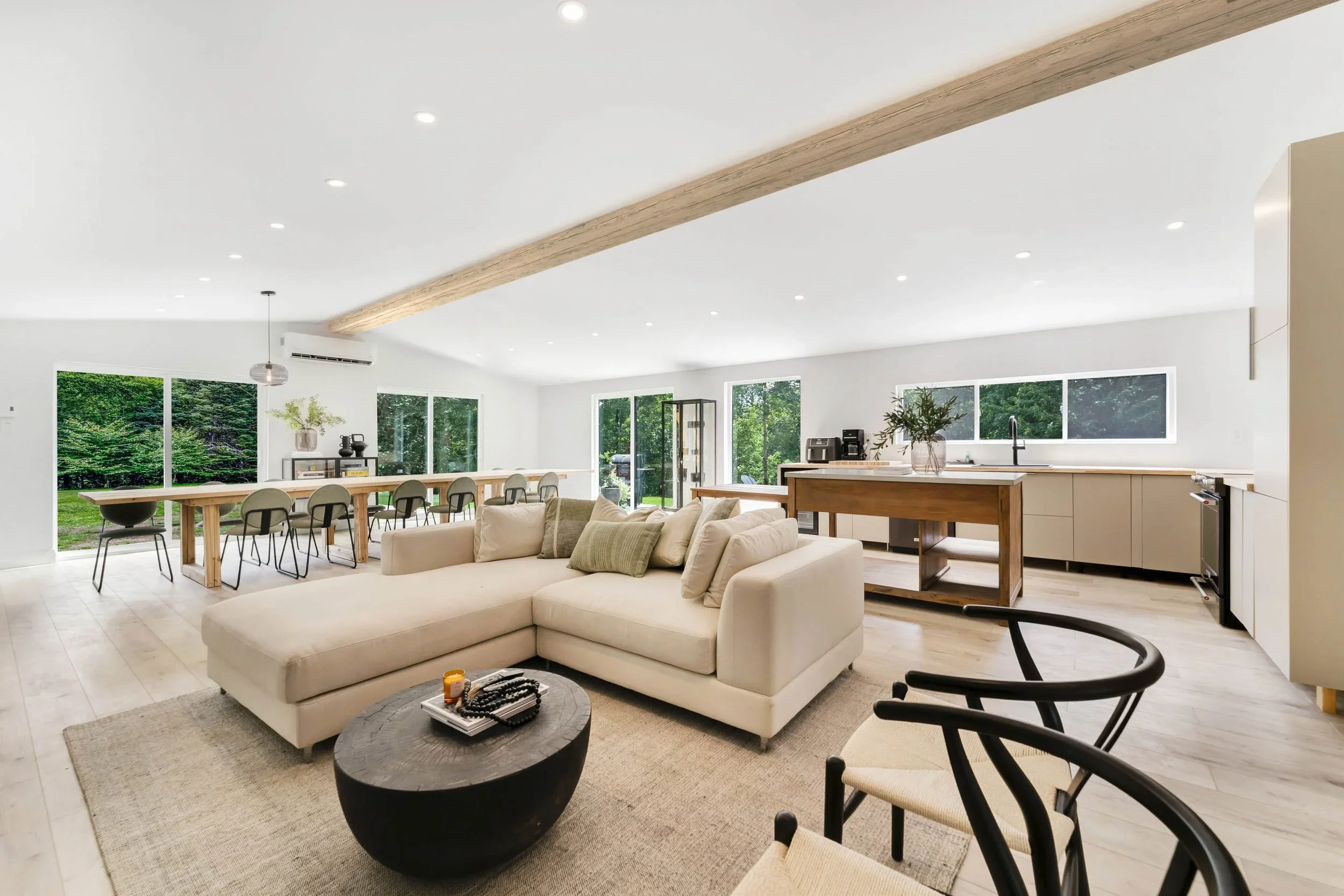 Intérieur lumineux et moderne d'une maison avec salon et cuisine ouverte, fenêtres panoramiques donnant sur un espace vert extérieur, mobilier beige et bois clair.