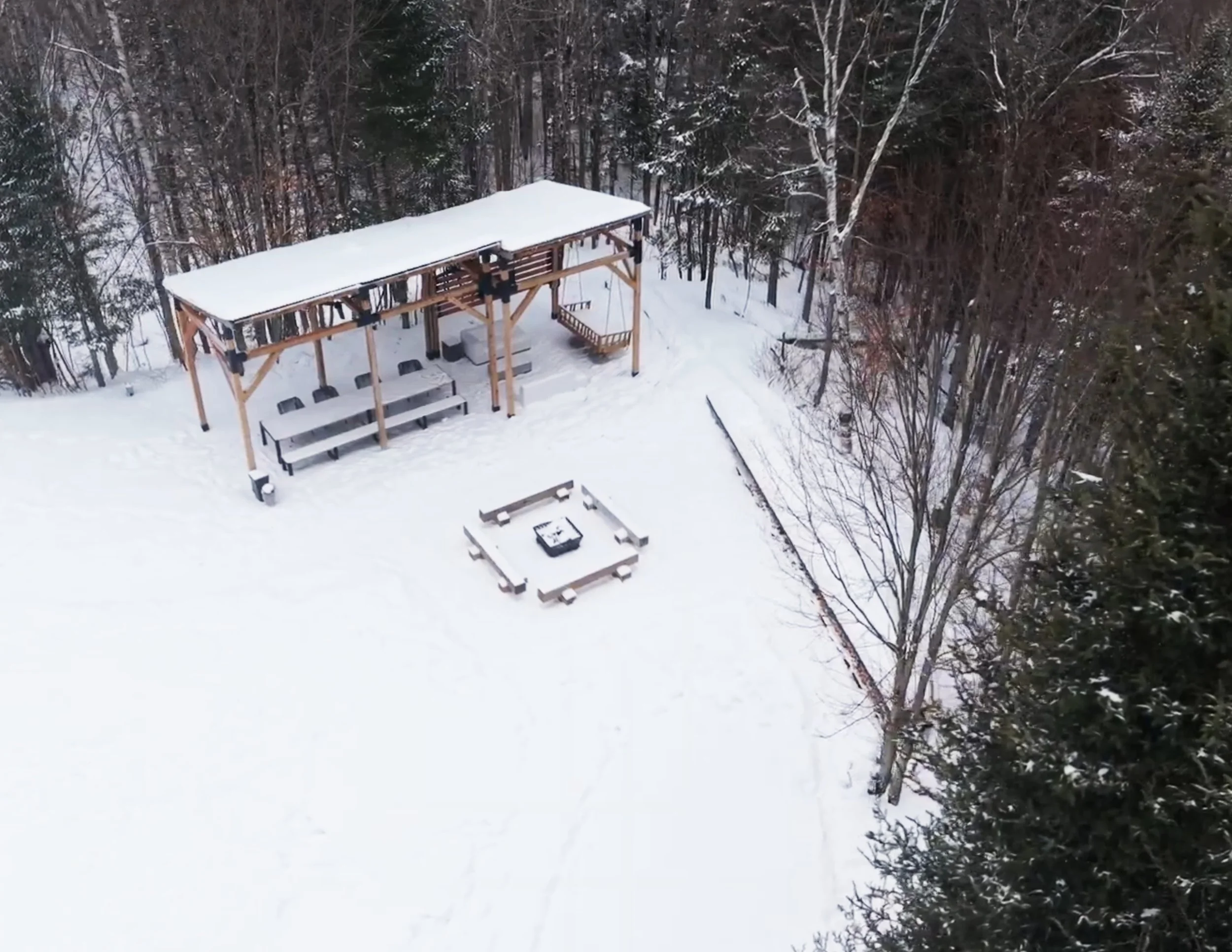 Vue aérienne du pavillon et du foyer extérieur sous la neige, entourés de forêt