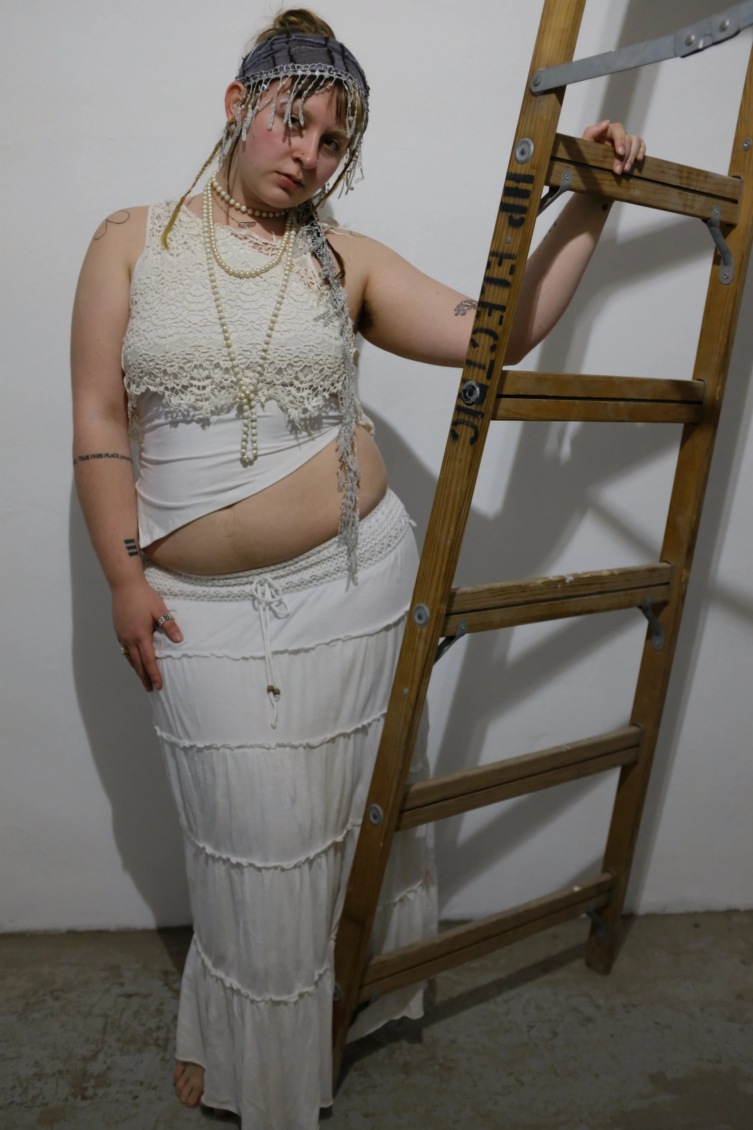 A woman with blonde dreadlocks and tattoos dressed in bohemian style, wearing a lace top, layered necklaces, and loose white pants, standing next to a wooden ladder against a plain white wall.