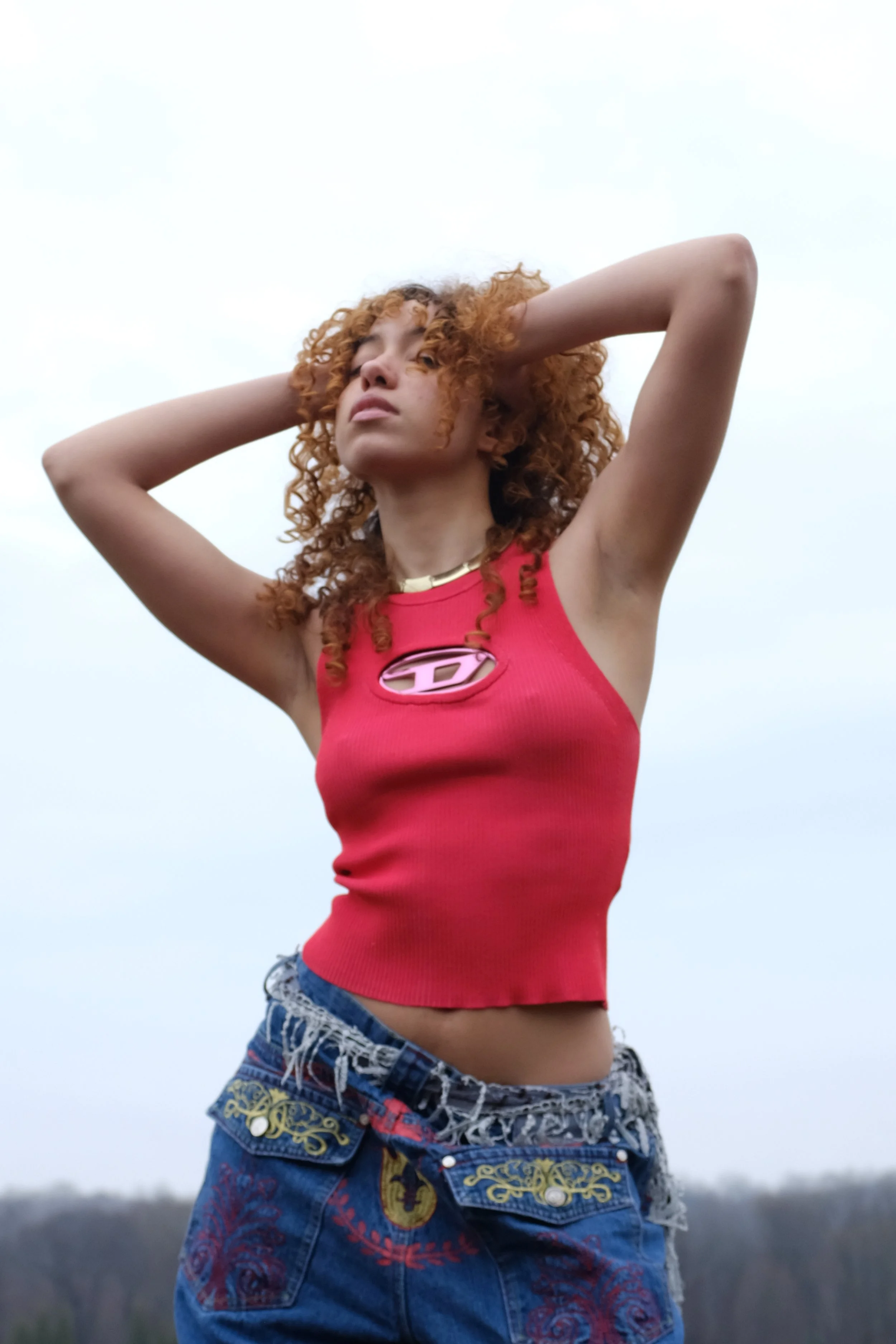 A young woman with curly red hair, wearing a red tank top and denim shorts with embroidered details, stands outdoors with her hands behind her head, gazing into the distance against a cloudy sky.