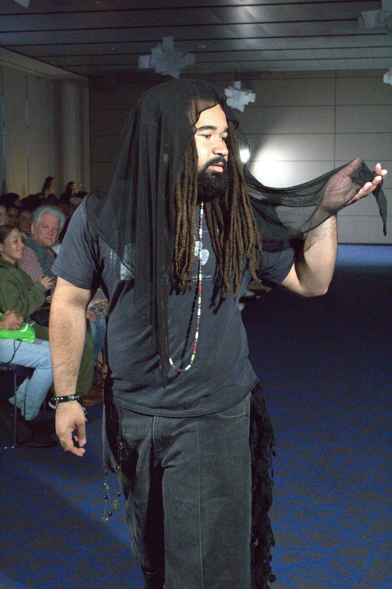 A man with long dreadlocks, a beard, and a black sheer cloth draped over his head and shoulder walking in an indoor setting with seated audience members watching.
