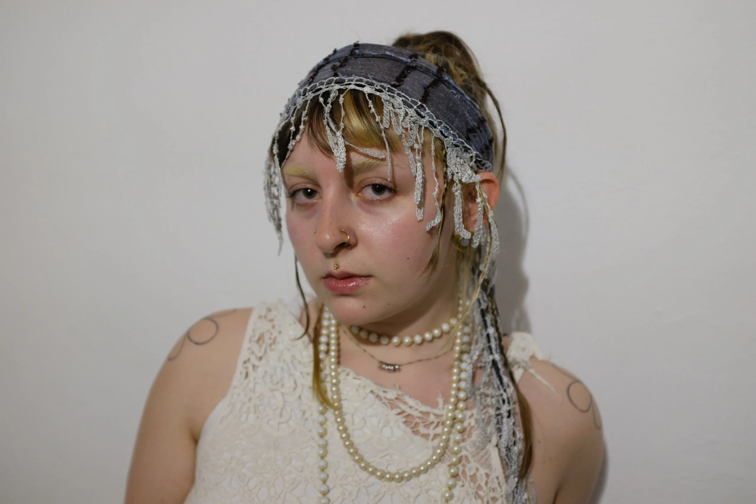 Young woman with light skin and multiple piercings, wearing layered pearl necklaces, a lace top, a headscarf with lace details, and tattoos on her shoulders, standing against a plain white wall.