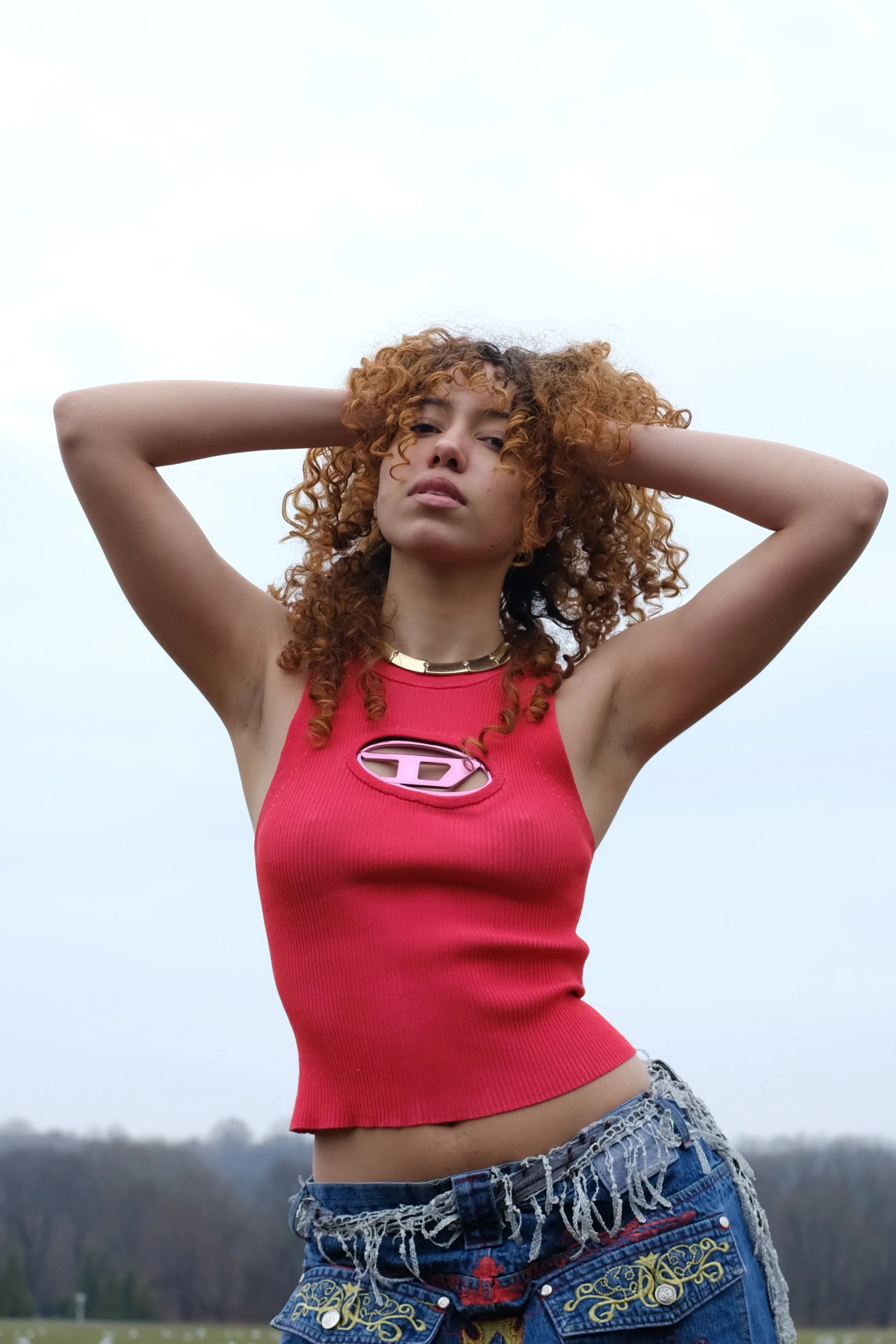 A young woman with curly red hair standing outdoors with her arms raised behind her head, wearing a red sleeveless top and distressed denim shorts.