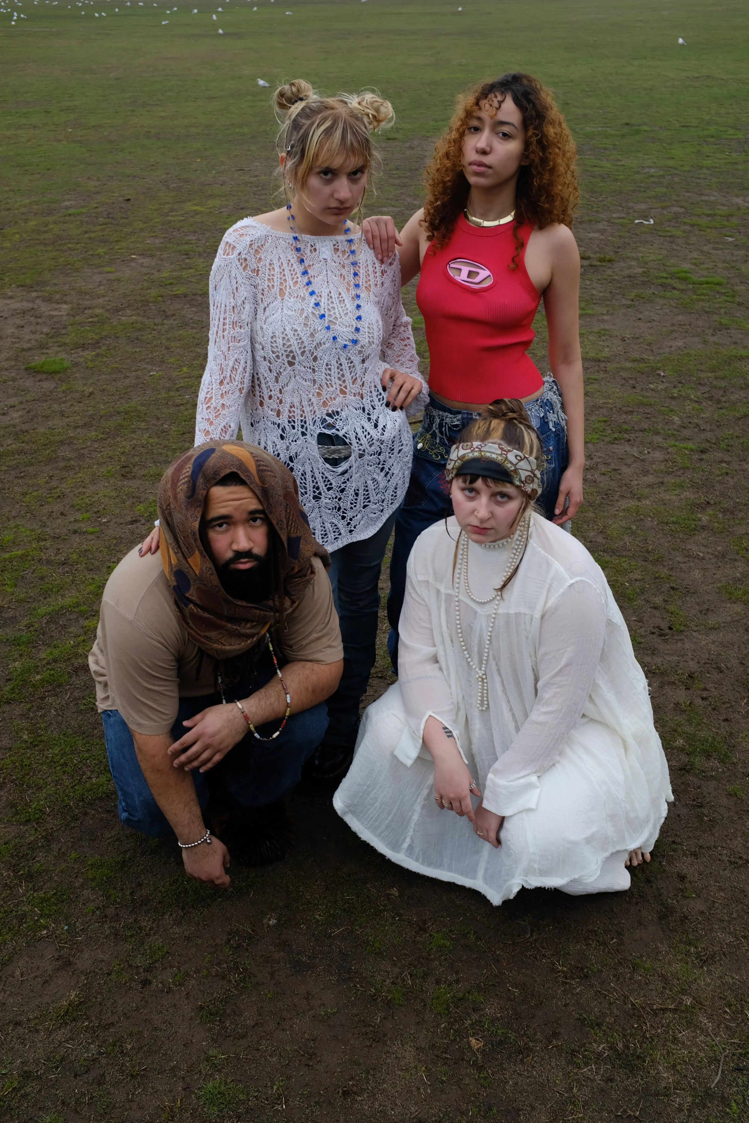 Group of five young adults outdoors on a muddy grassy field, some crouching and some standing, dressed in bohemian and casual streetwear styles.