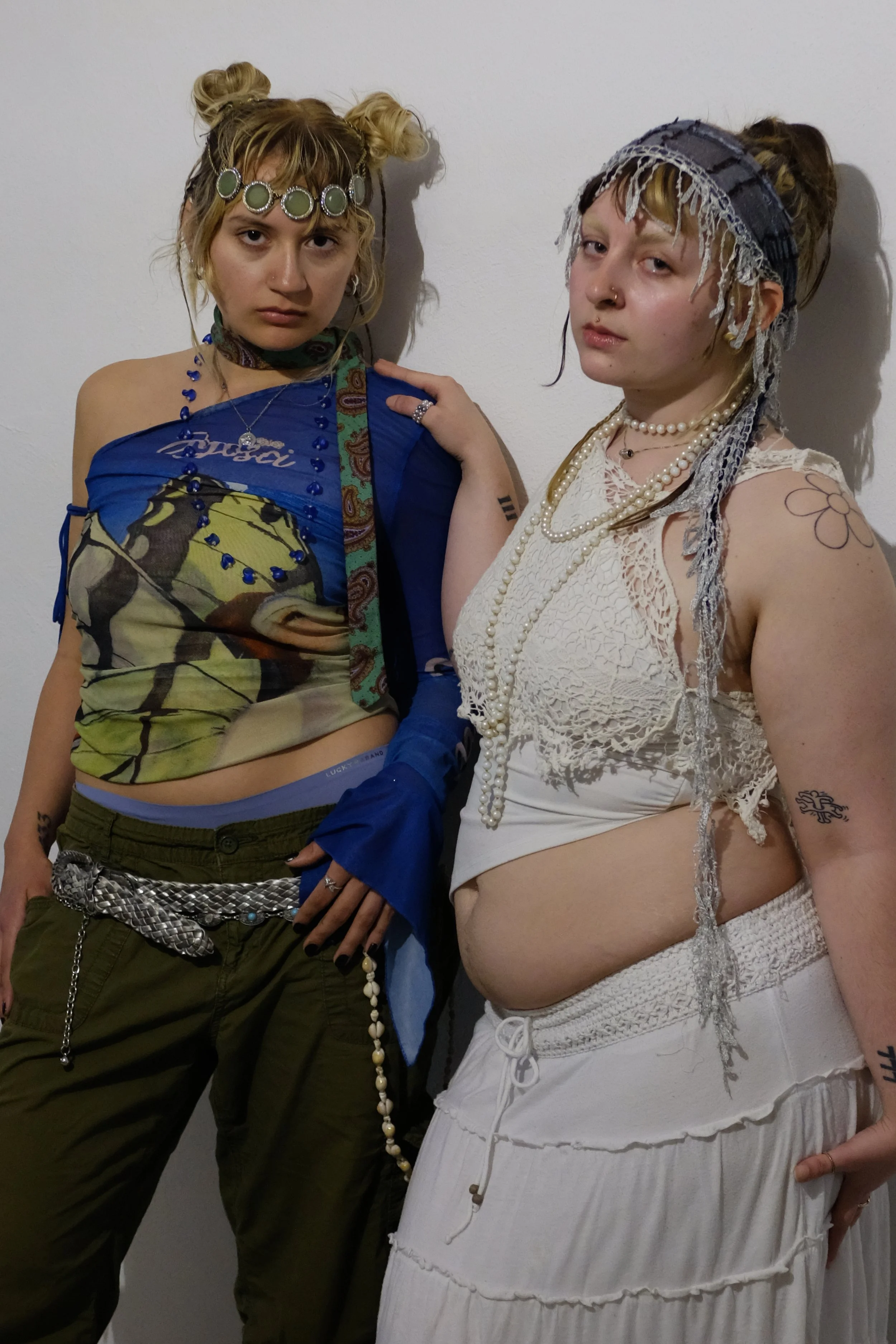 Two young women with tattoos and styled hair posing against a plain white wall. They wear bohemian and eclectic clothing and accessories, including jewelry, headbands, and layered necklaces.