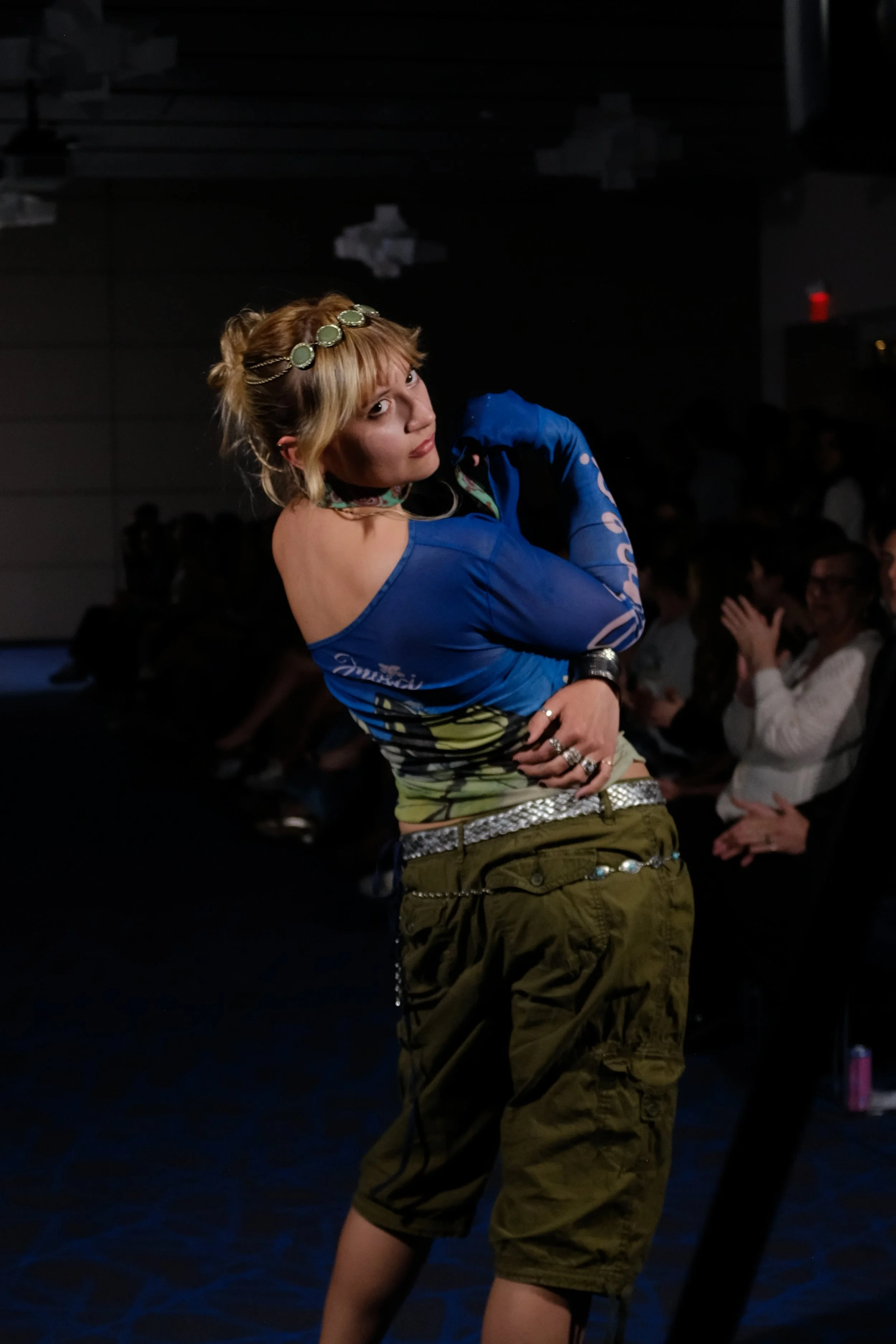 A woman with blonde hair styled in a messy bun, wearing a green headband, a blue off-shoulder top, and olive green cargo shorts, striking a pose at a fashion event with an audience clapping in the background.