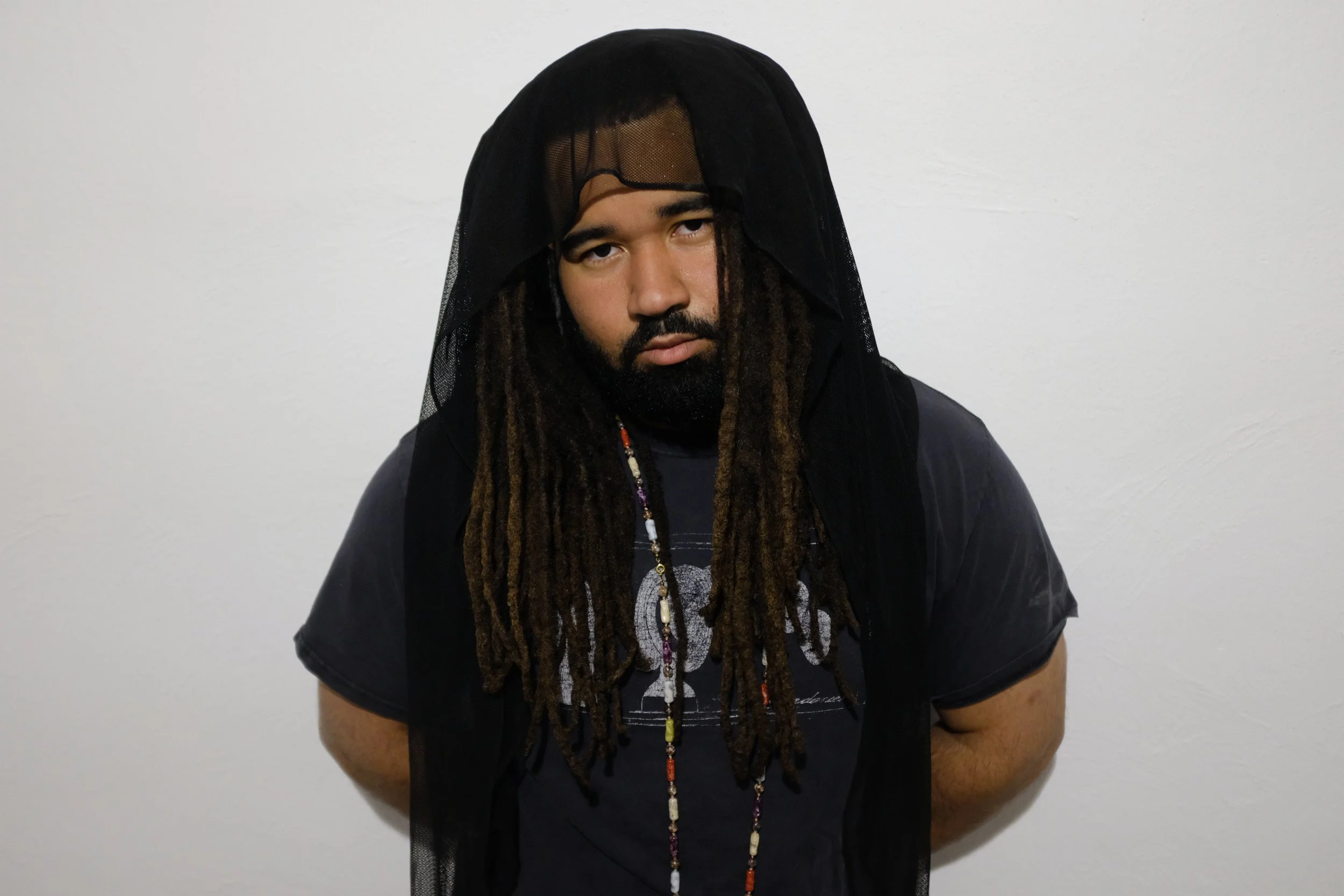 A man with dreadlocks wearing a black t-shirt and a black veil over his head stands in front of a plain white wall.
