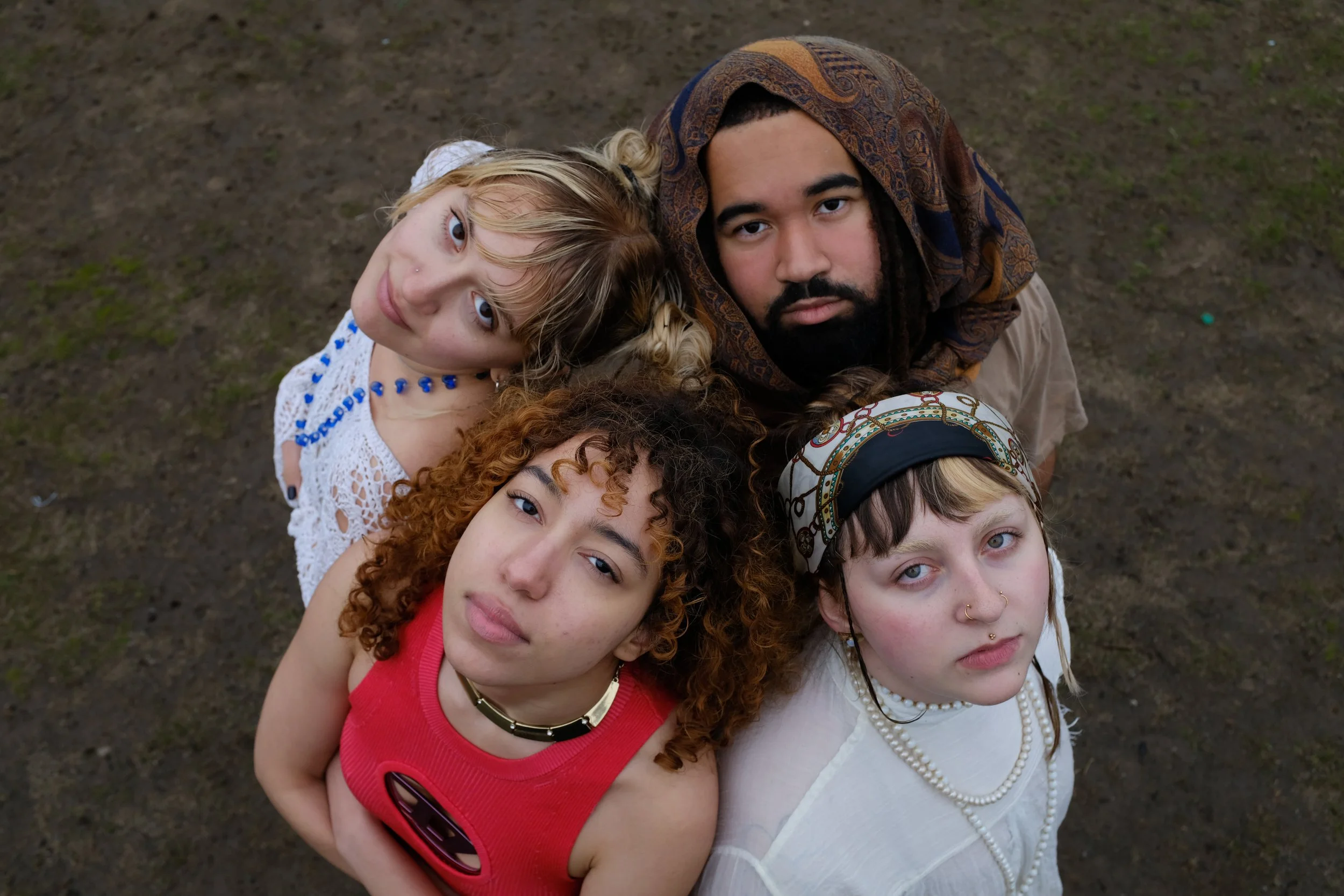 A group of five young women and one young man standing closely together outdoors, looking up at the camera. They have diverse hair colors and styles, and are wearing casual and unique clothing, including jewelry and accessories.