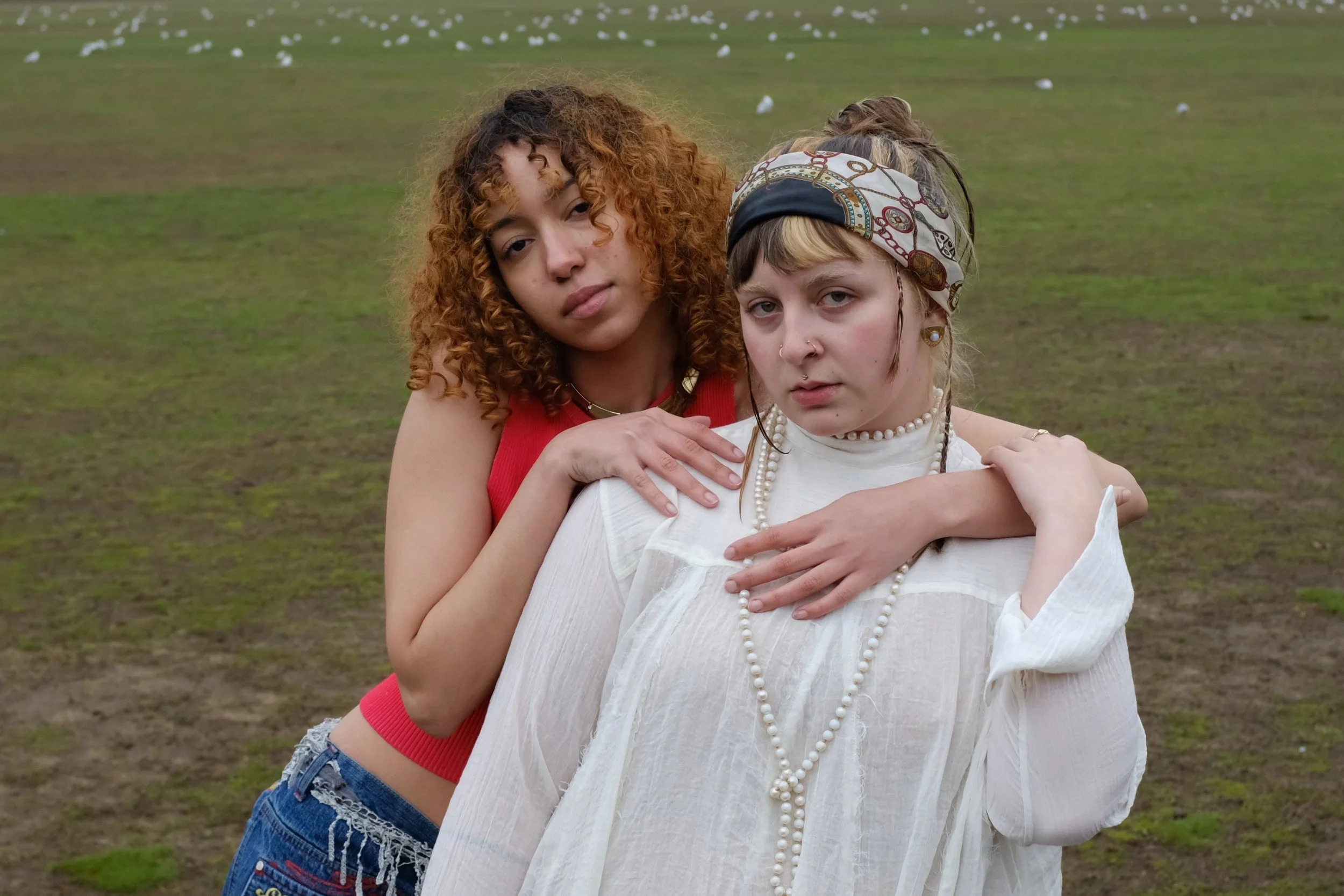 Two young women with curly and straight hair, one with a bandana, standing in a field with seagulls flying above.