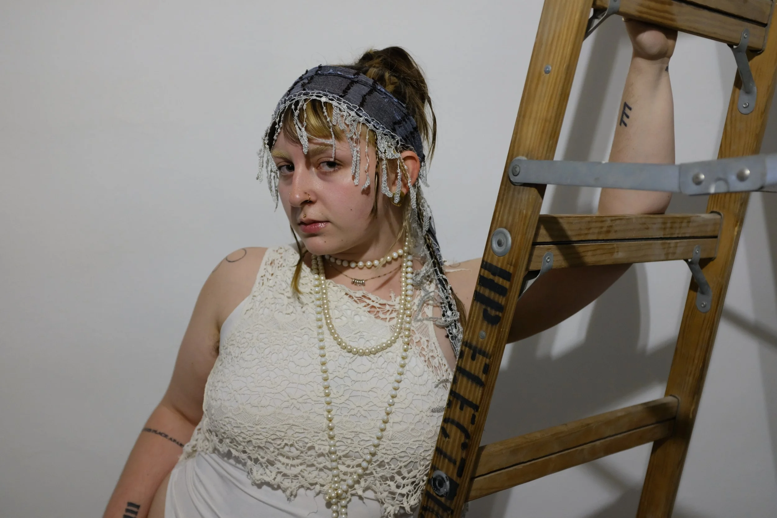 A woman with tattoos, jewelry, and headscarf leaning on a wooden ladder against a plain white wall.
