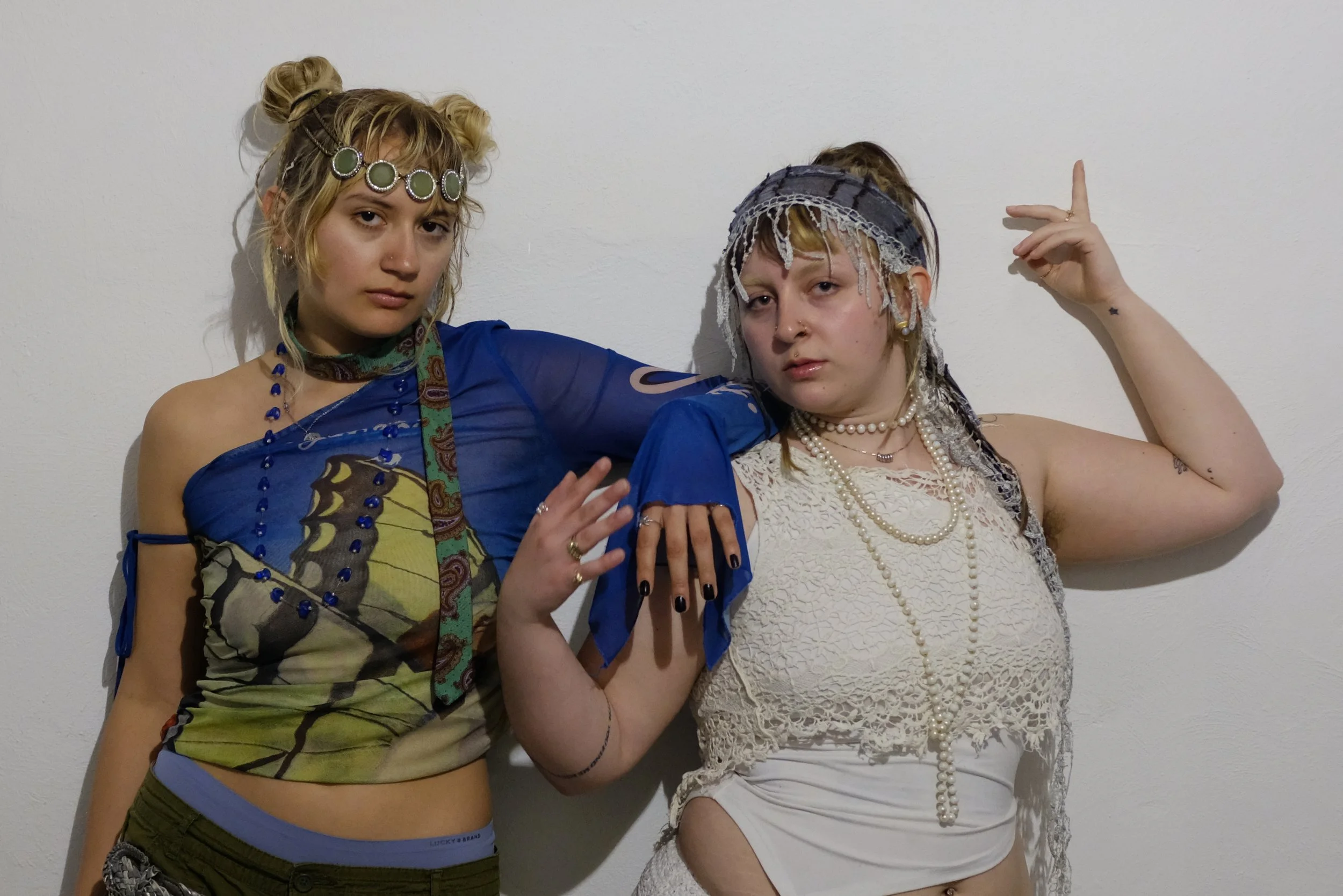 Two young women posing against a plain white wall, dressed in eclectic, bohemian style outfits with accessories, jewelry, and headpieces.