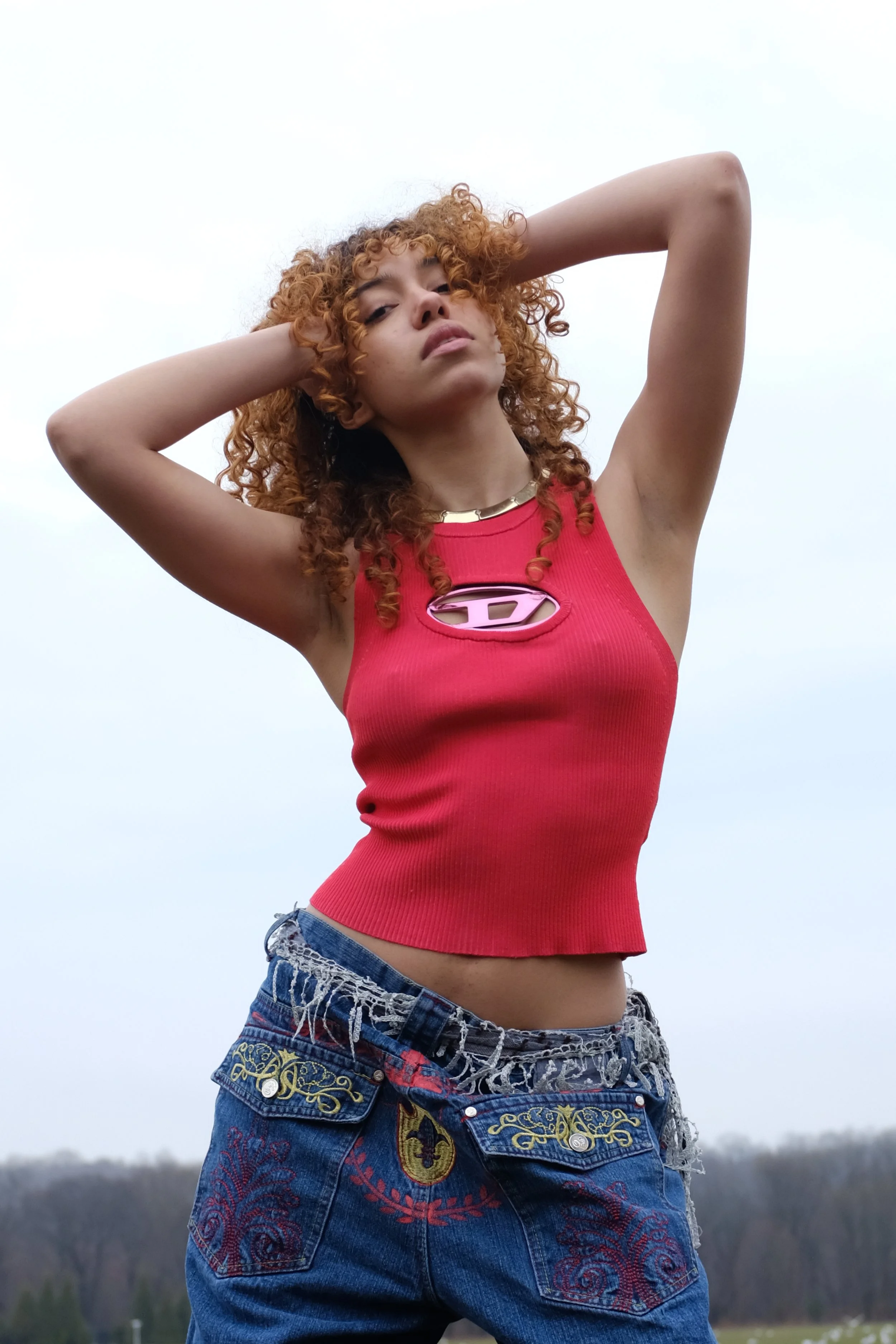 Young woman with curly red hair posing outdoors against a gray sky, wearing a red sleeveless top with a Superman logo and decorated blue jeans.