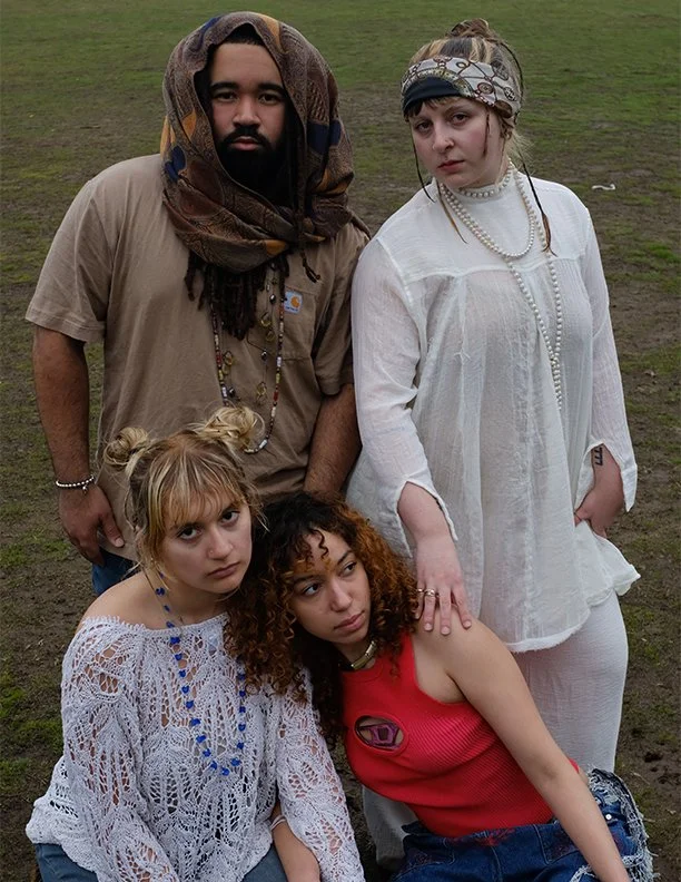 A group of four diverse young adults standing and sitting outdoors on grass, dressed in casual and vintage-style clothing, with serious expressions.