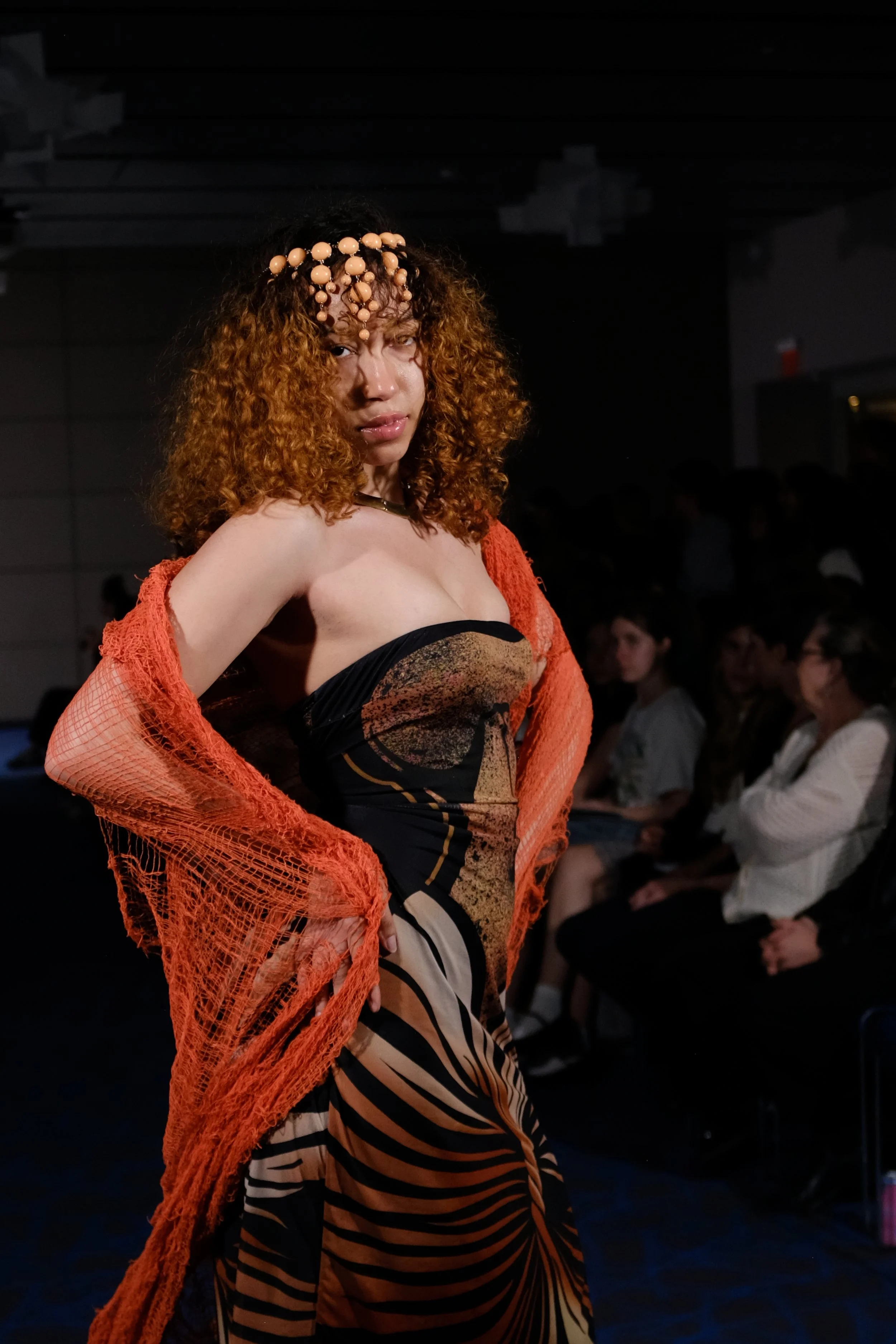 Fashion model walking on runway during a fashion show, wearing a strapless dress with a spiral pattern and a sheer orange shawl, with audience in background.