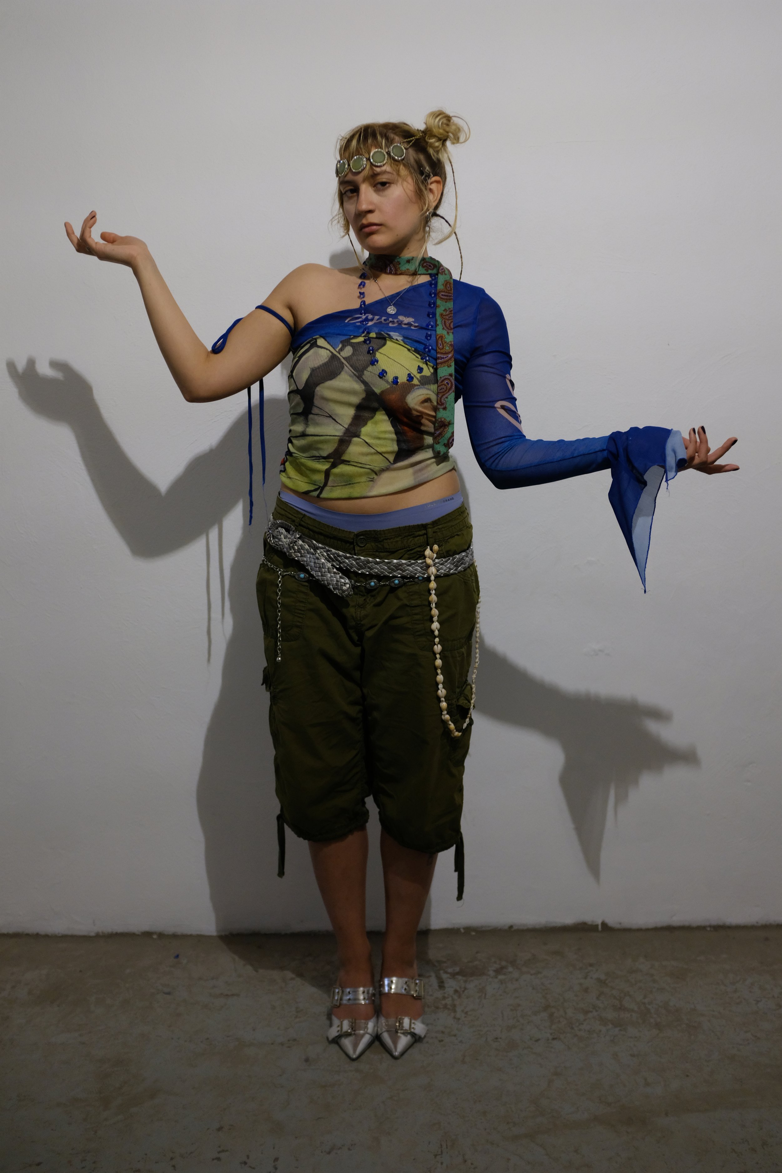 A woman standing against a white wall, dressed in a colorful, eclectic outfit with layered necklaces, green pants, pointed silver shoes, and a headband, with her armsoutrstretched and a neutral expression.