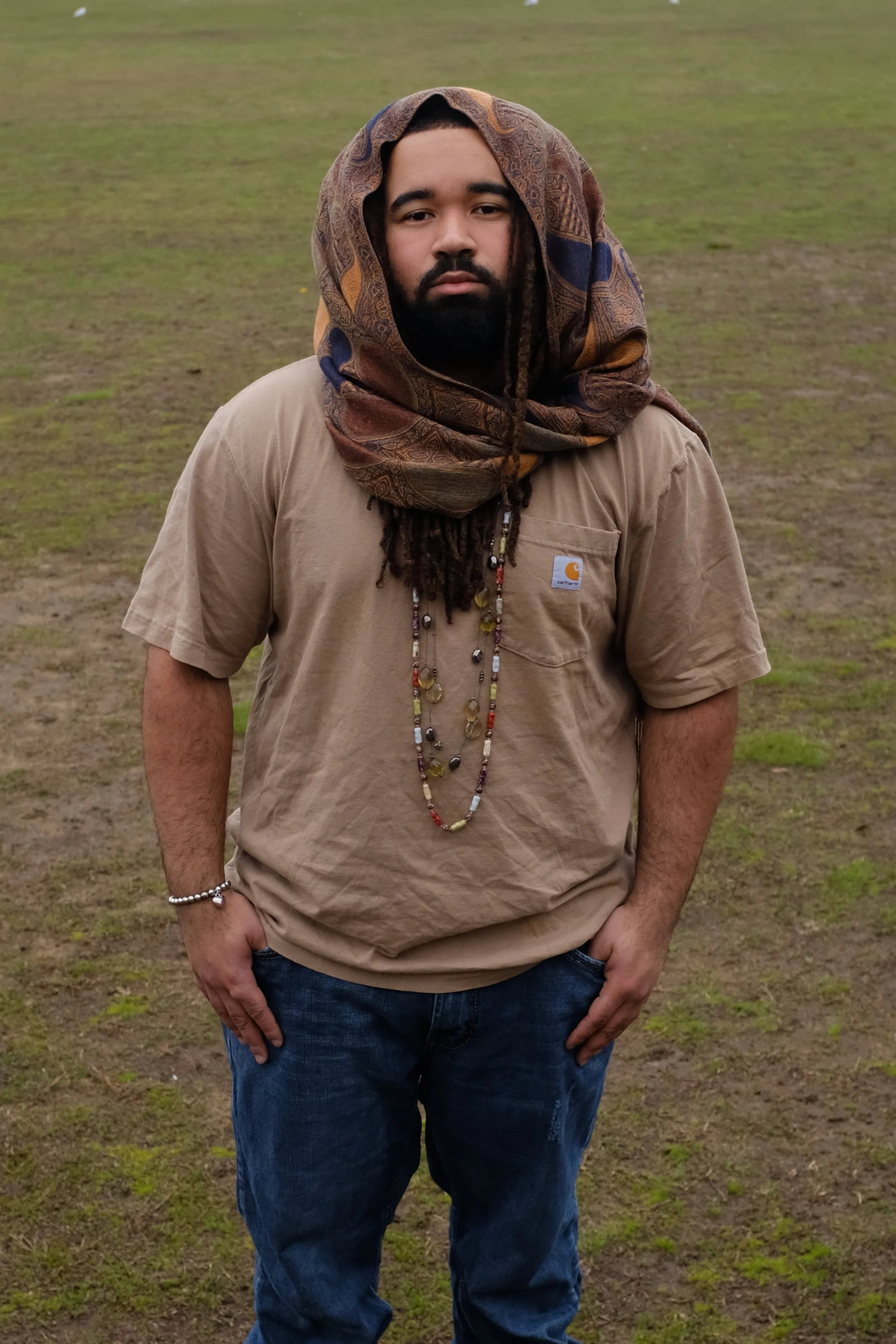 A man standing outdoors on a grassy area, wearing a beige Carhartt shirt, jeans, and a multicolored scarf wrapped around his head, with long necklaces, a silver bracelet, and beaded hair.