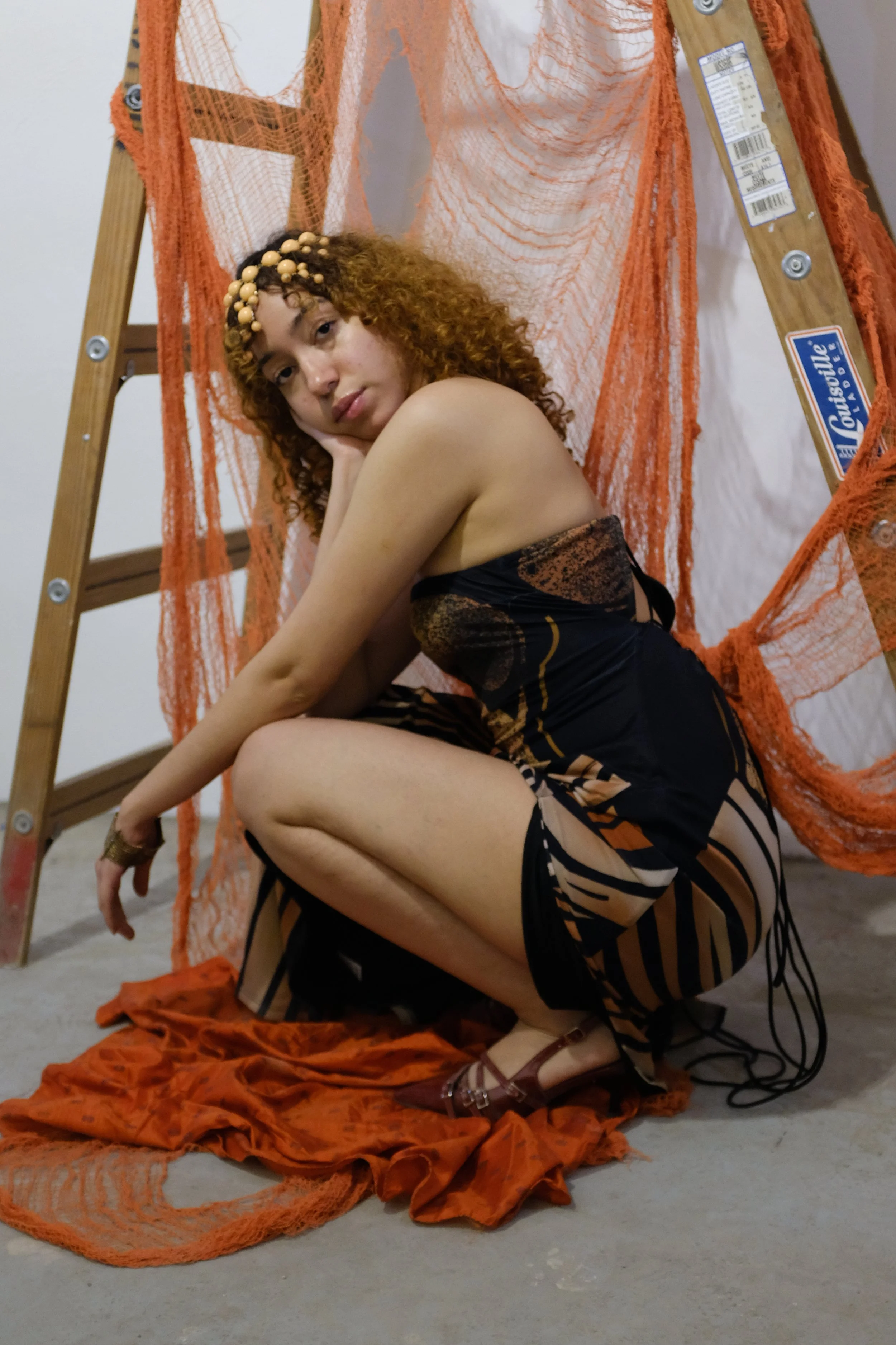A woman with curly red hair, wearing a patterned dress and high heels, squats on the floor with a contemplative expression, surrounded by orange nets and fabric, next to a wooden ladder.