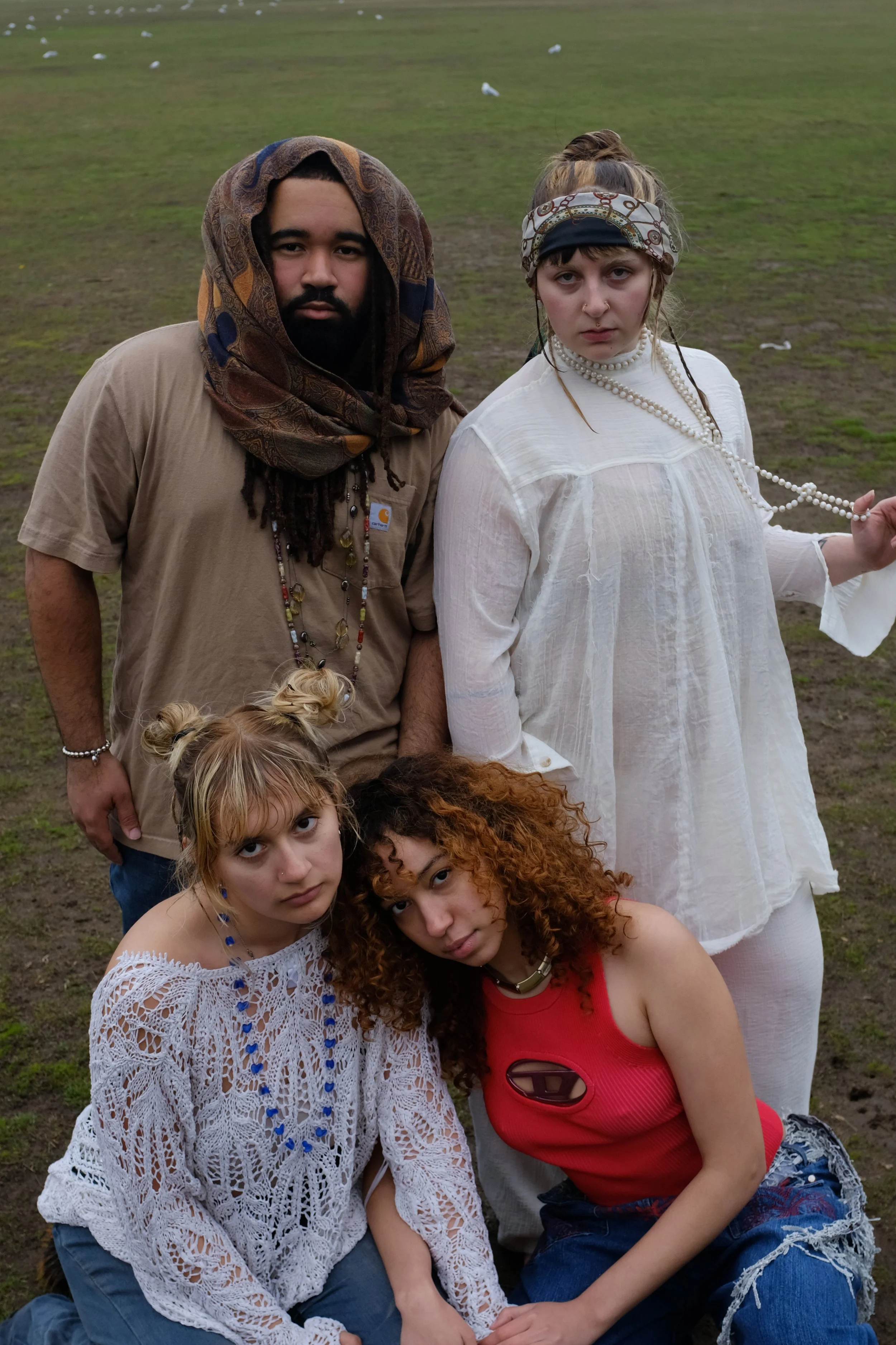 A group of five young people posing outdoors in a grassy area, with serious expressions and casual bohemian style clothing and accessories.