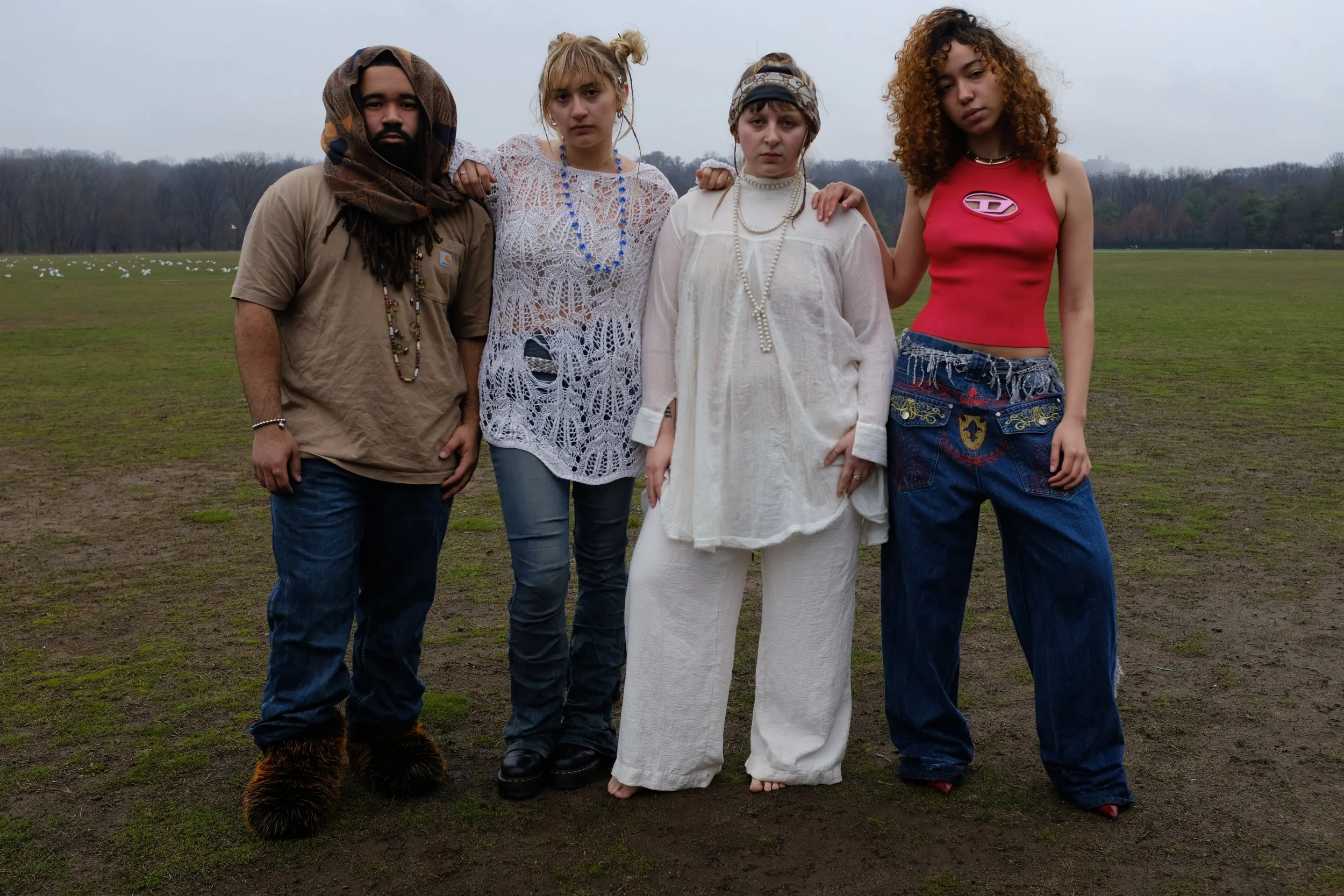 Four young adults standing together outdoors on a cloudy day with a grassy field and trees in the background. They are dressed in eclectic, bohemian clothing.