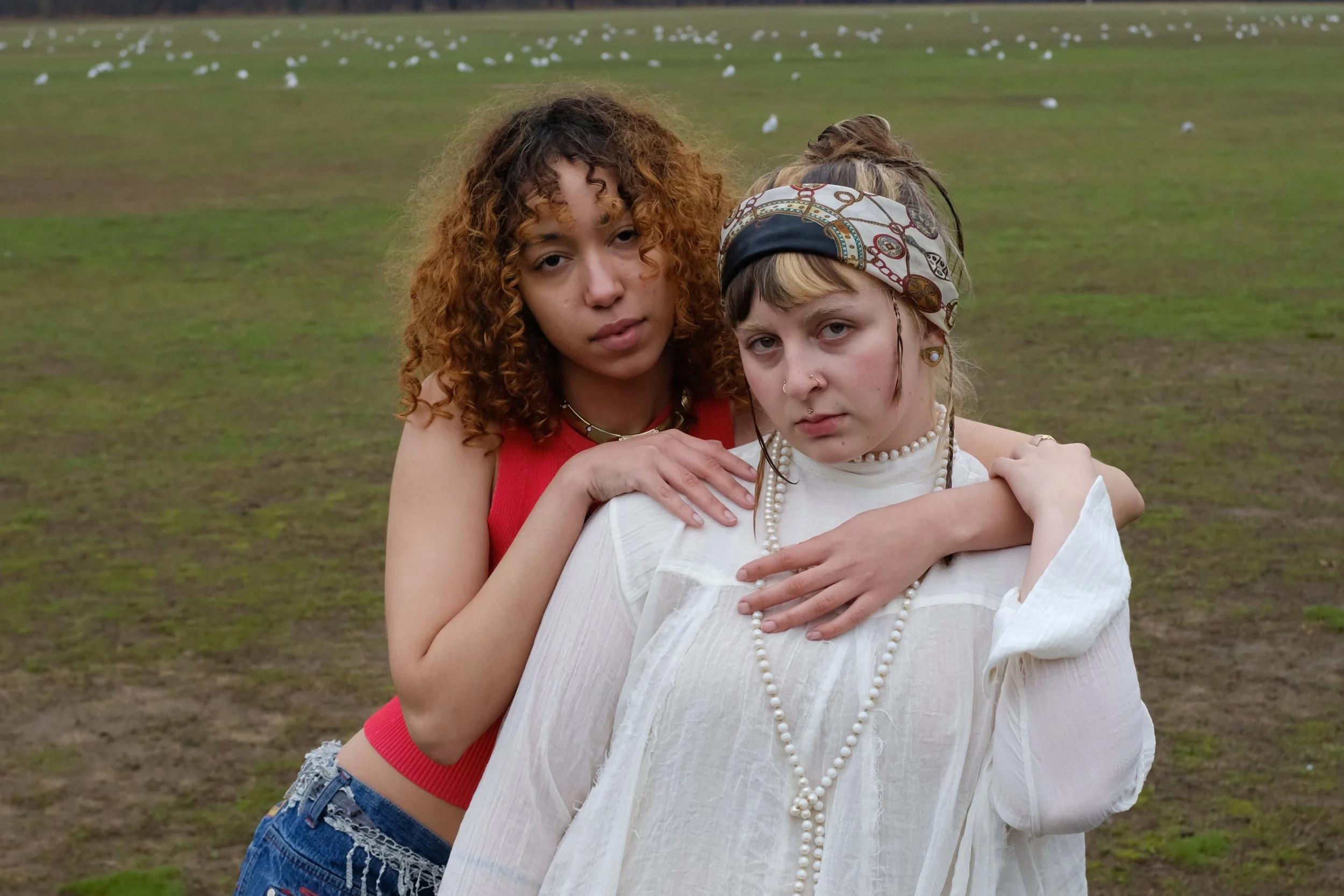 Two young women standing outdoors with a grassy field and seagulls in the background, one in a red top and denim shorts, the other in a white blouse with pearl accessories and a headscarf.