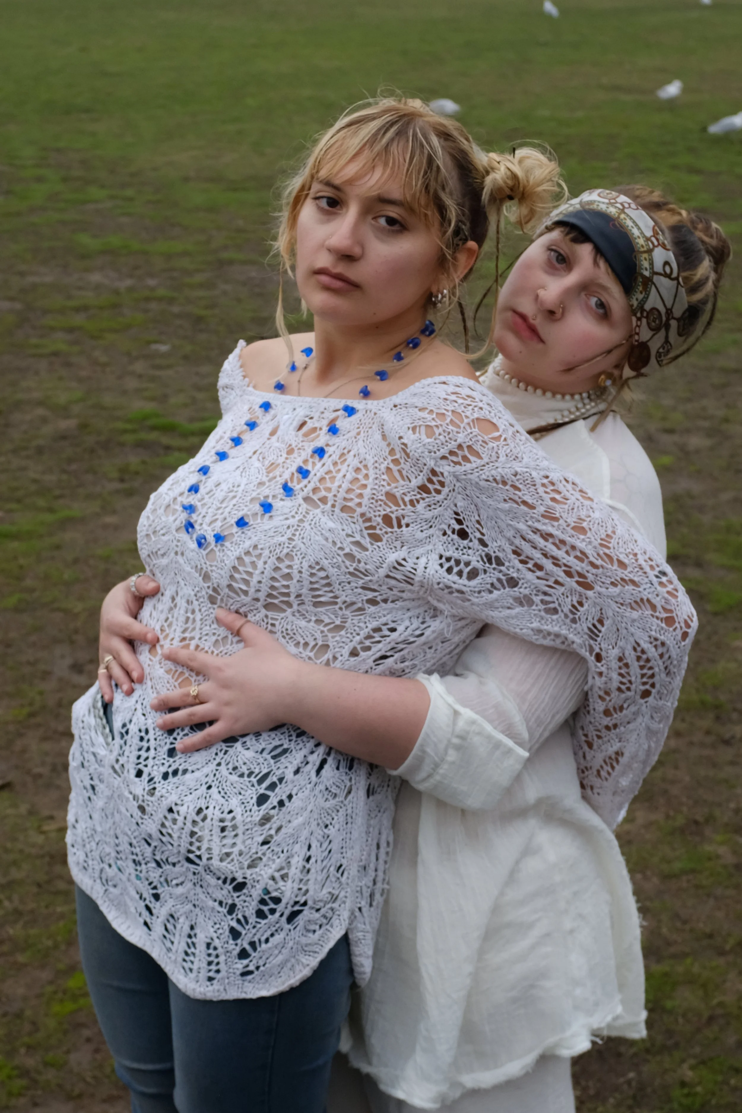 Two young women pose closely together outdoors, one in front with a lace white top and blue jewelry, the other behind with a headscarf, pearl necklace, and white clothing, against a grassy background with seagulls.