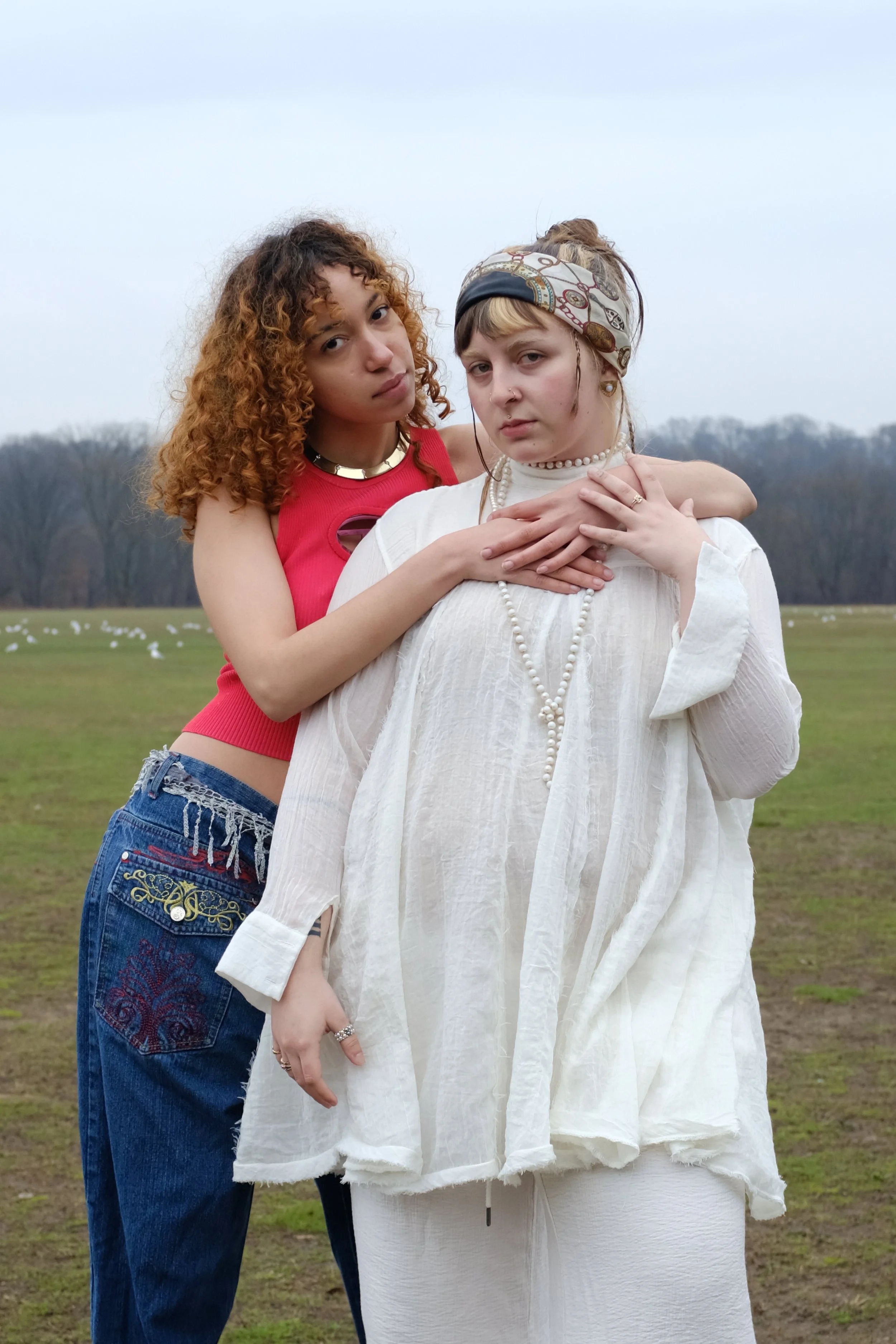 Two young women standing outdoors in a grassy field with trees in the background. One has curly red hair and is wearing a pink crop top, and the other has light brown hair with a headscarf and is dressed in white, looking serious while embracing each
